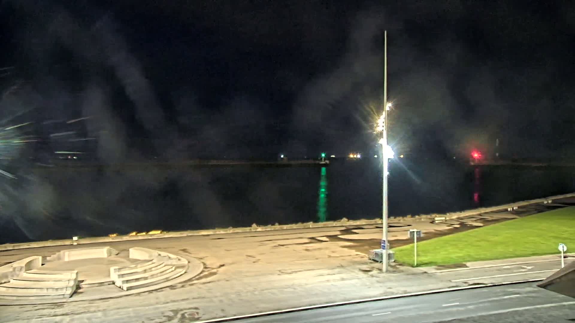 Fireworks explode over a dark body of water at night, illuminating a concrete amphitheater and nearby roadway.