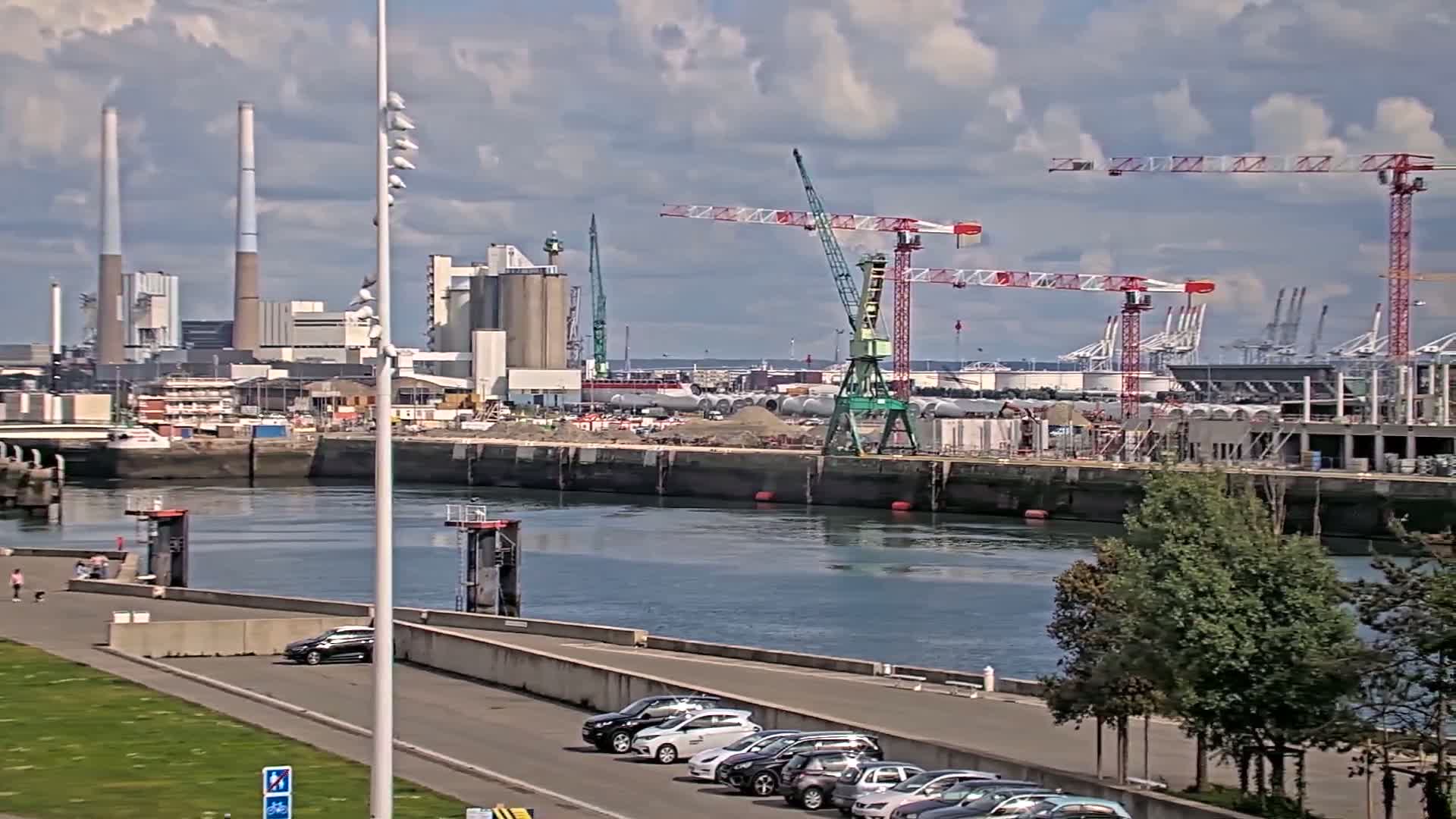 A partly cloudy day shows a waterway with parked cars, a grassy area, and an industrial area across the water with cranes and smokestacks.