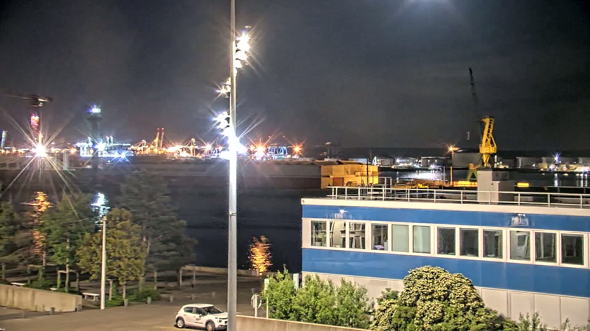 A nighttime view of a brightly lit industrial harbor across a body of water, with a small car parked in a lot in the foreground, near a blue building.