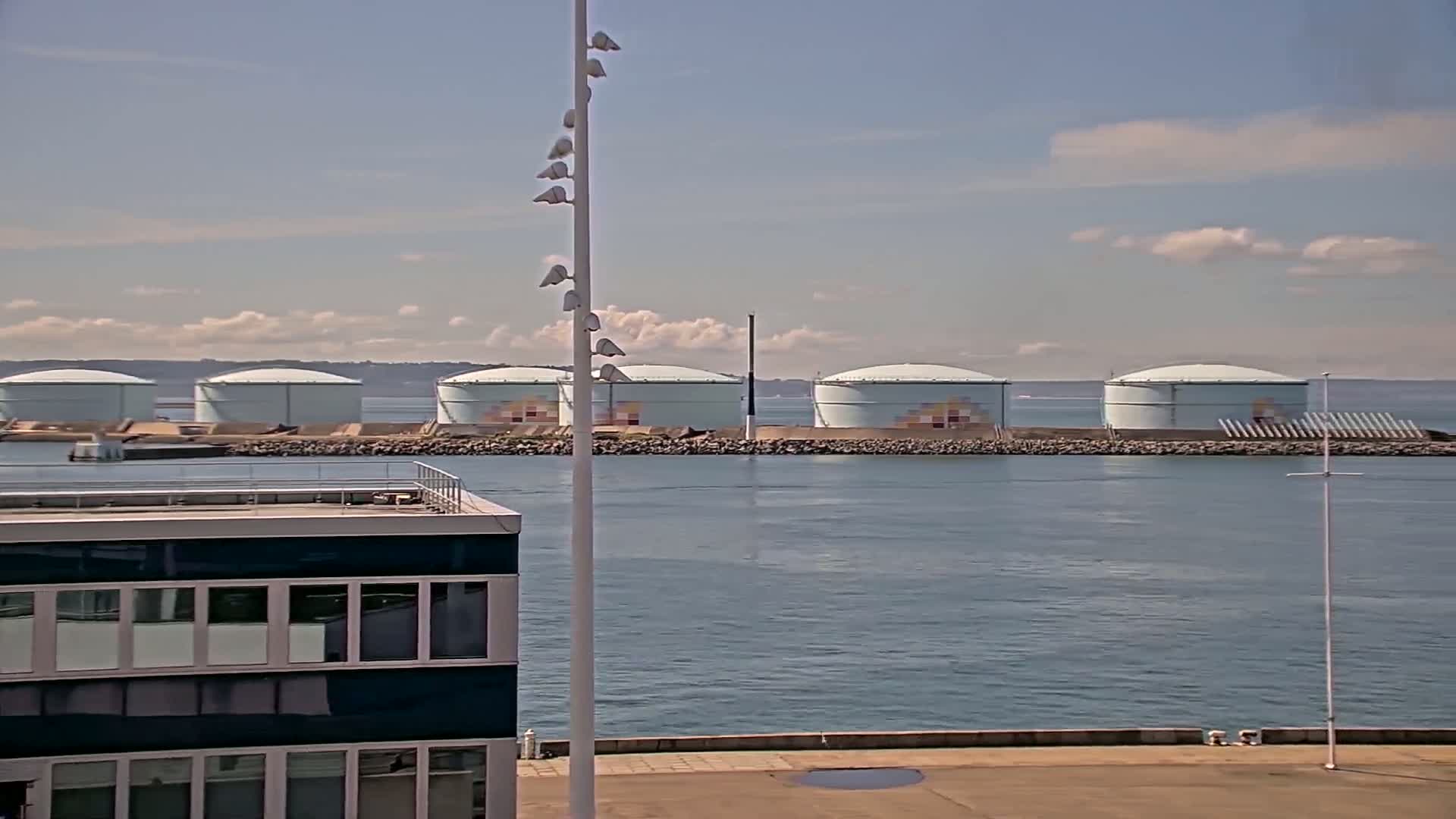 Several large, light-blue storage tanks sit on a breakwater in calm water under a mostly sunny sky.
