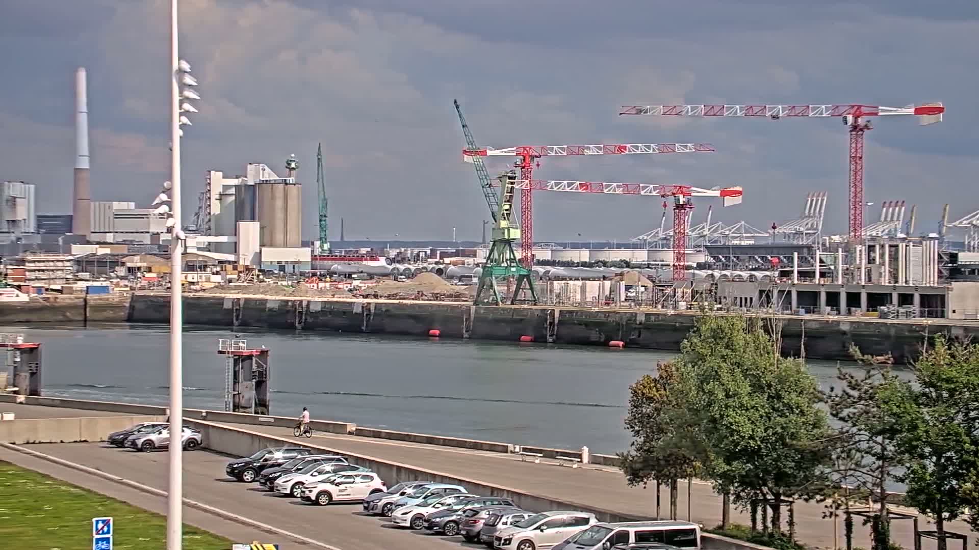A waterfront construction site with several cranes, industrial buildings, and parked cars under a partly cloudy sky.