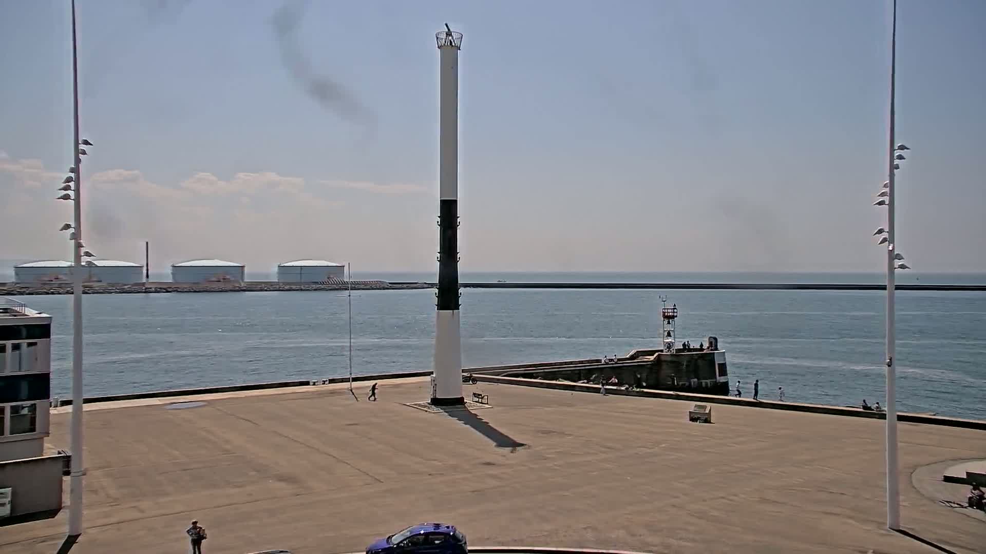 A tall, black and white lighthouse stands in a harbor on a sunny day, with several people and vehicles visible on a paved area near the water's edge, and large storage tanks in the distance.