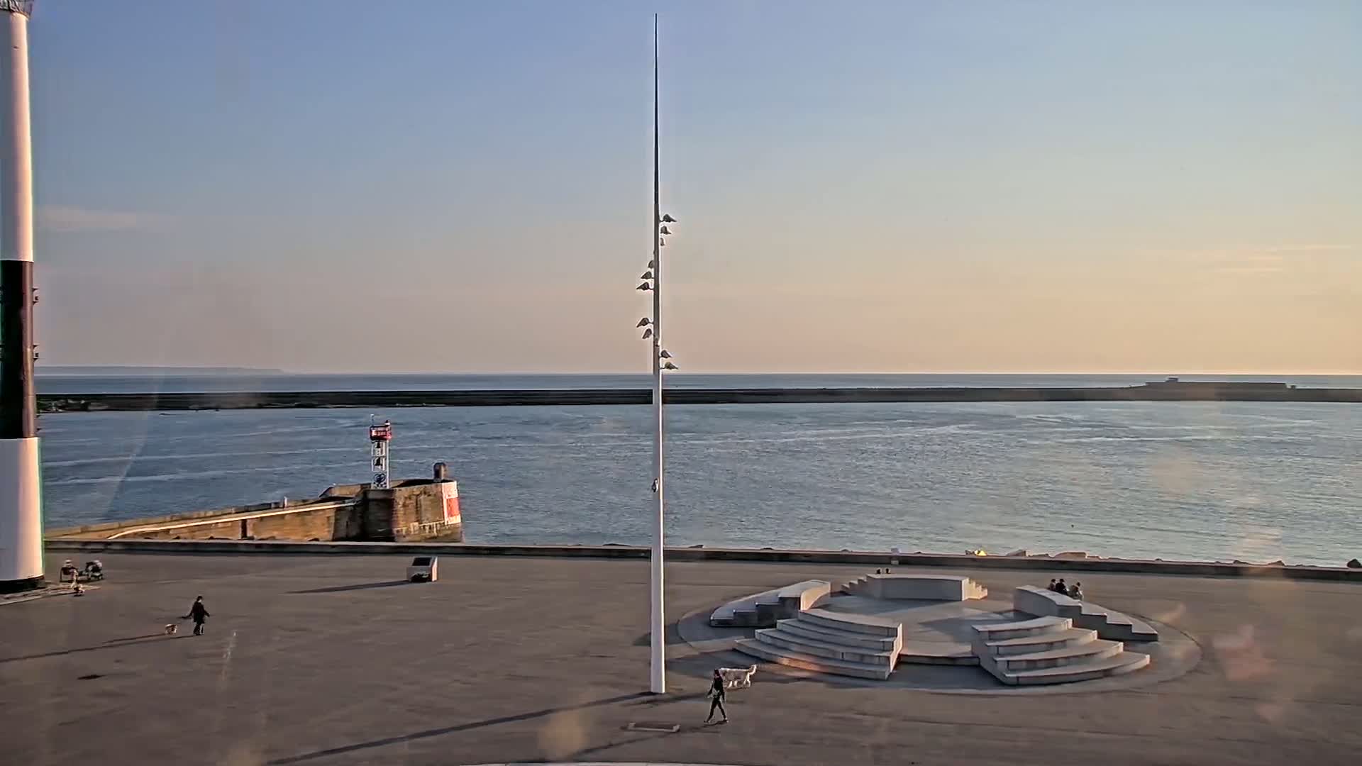 A mostly clear, calm day overlooks a harbor with a lighthouse, a tall pole with lights, a stone amphitheater, and several people walking on a paved area.