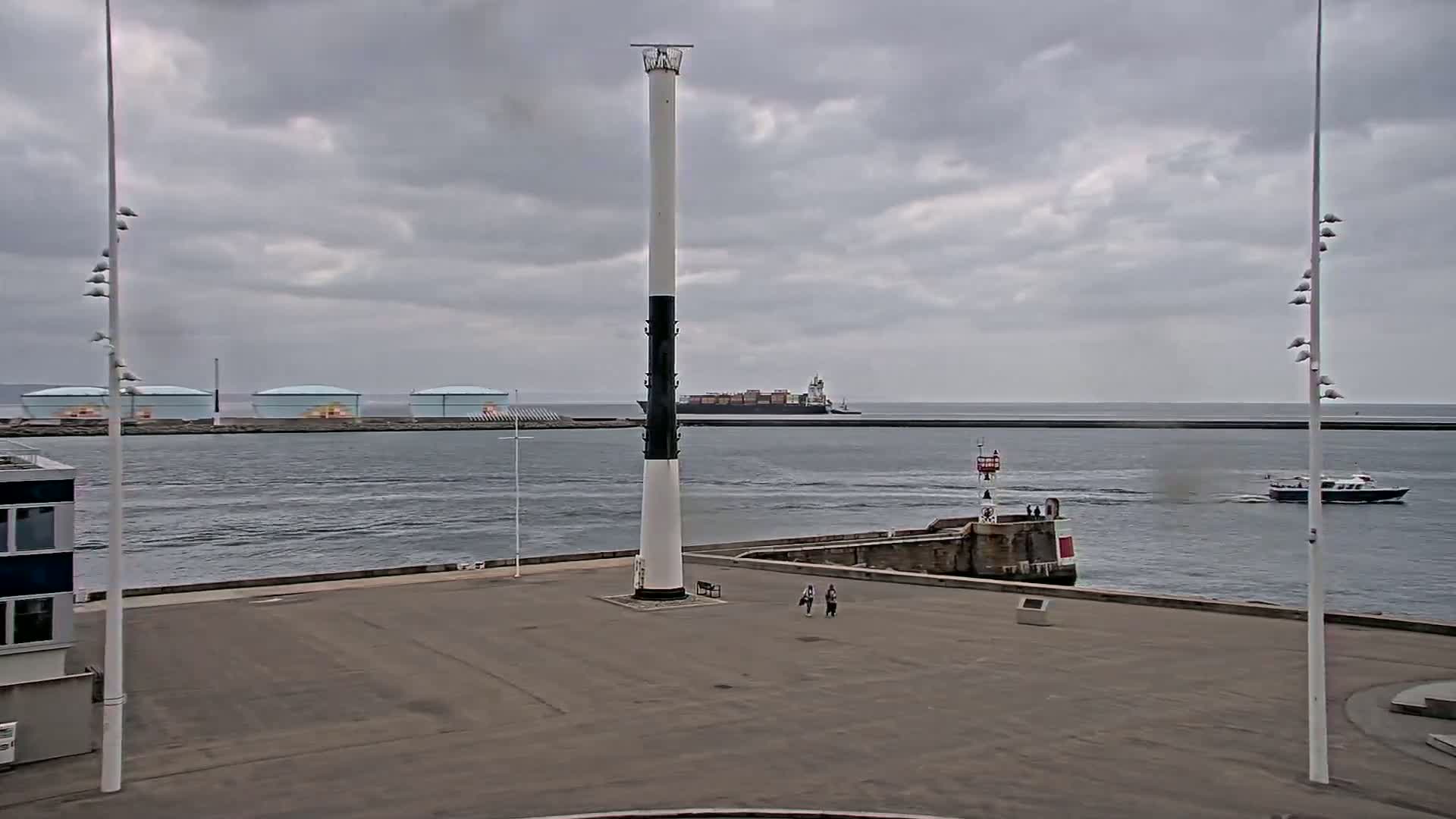 A tall, black-and-white striped pole stands in a harbor square under a cloudy sky, with a container ship and a small boat visible on the water, and several storage tanks in the distance.
