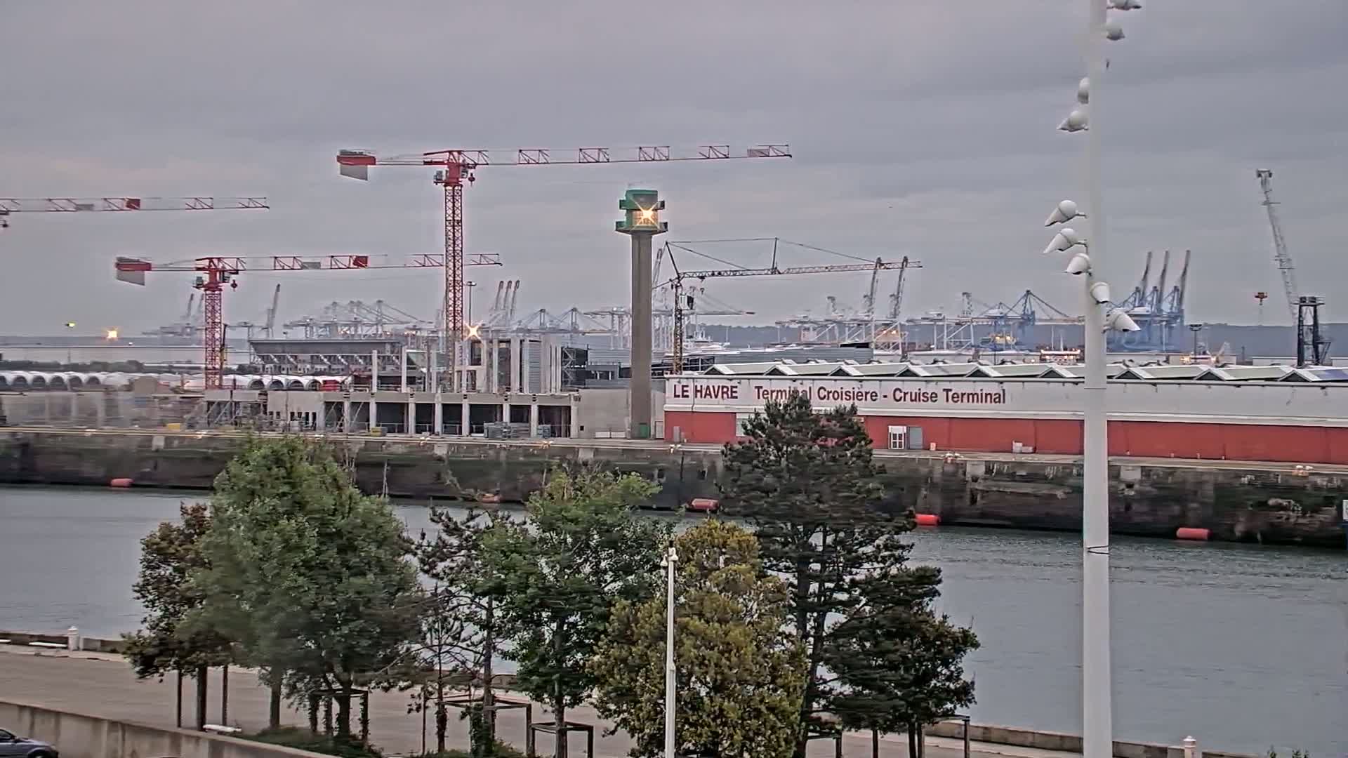 A harbor scene under an overcast sky shows a partially-constructed building near a cruise terminal, several cranes, and numerous ships beyond.