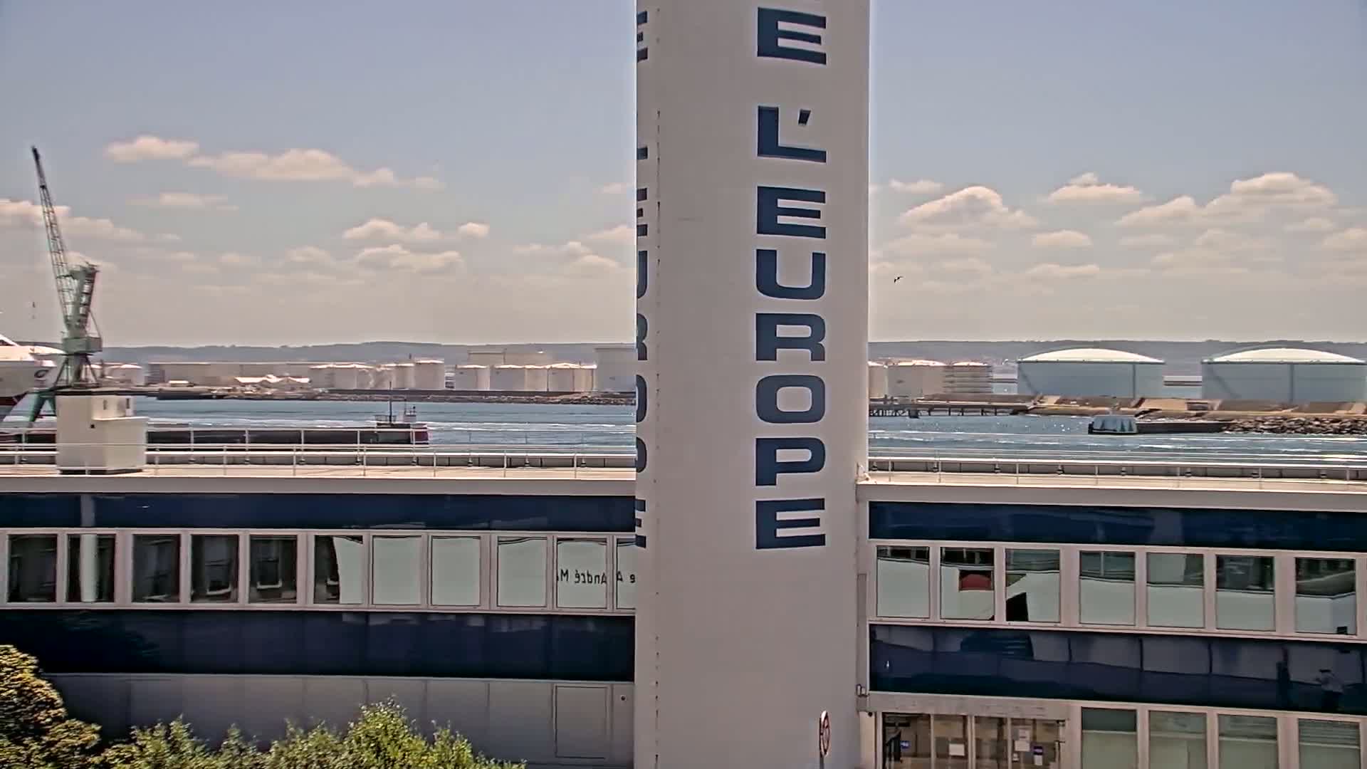 A partly cloudy day shows a large white tower with "L'Europe" written on it, next to a building with many windows overlooking a body of water with a distant industrial area and several storage tanks.
