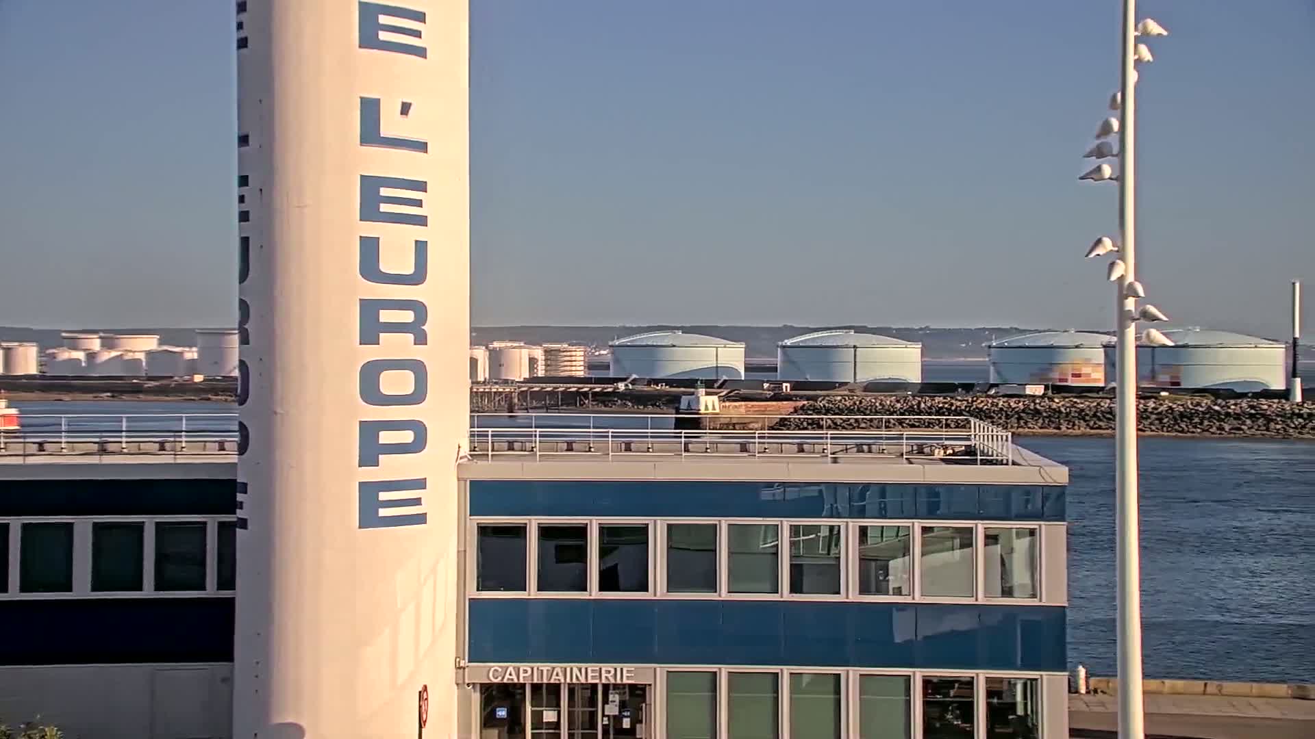 A large, white cylinder with blue lettering stands beside a blue building overlooking a body of water and several large industrial storage tanks under a clear, sunny sky.