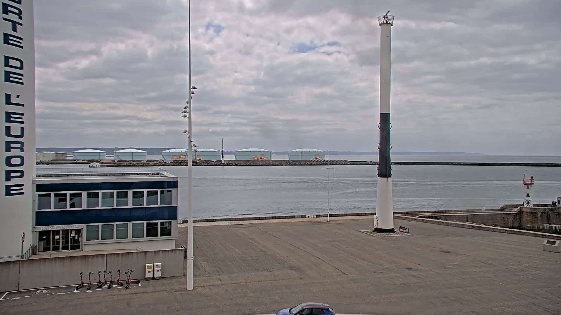 A harbor scene under a cloudy sky shows a tall, slender lighthouse, several large storage tanks in the distance, and a small building with electric scooters parked outside.