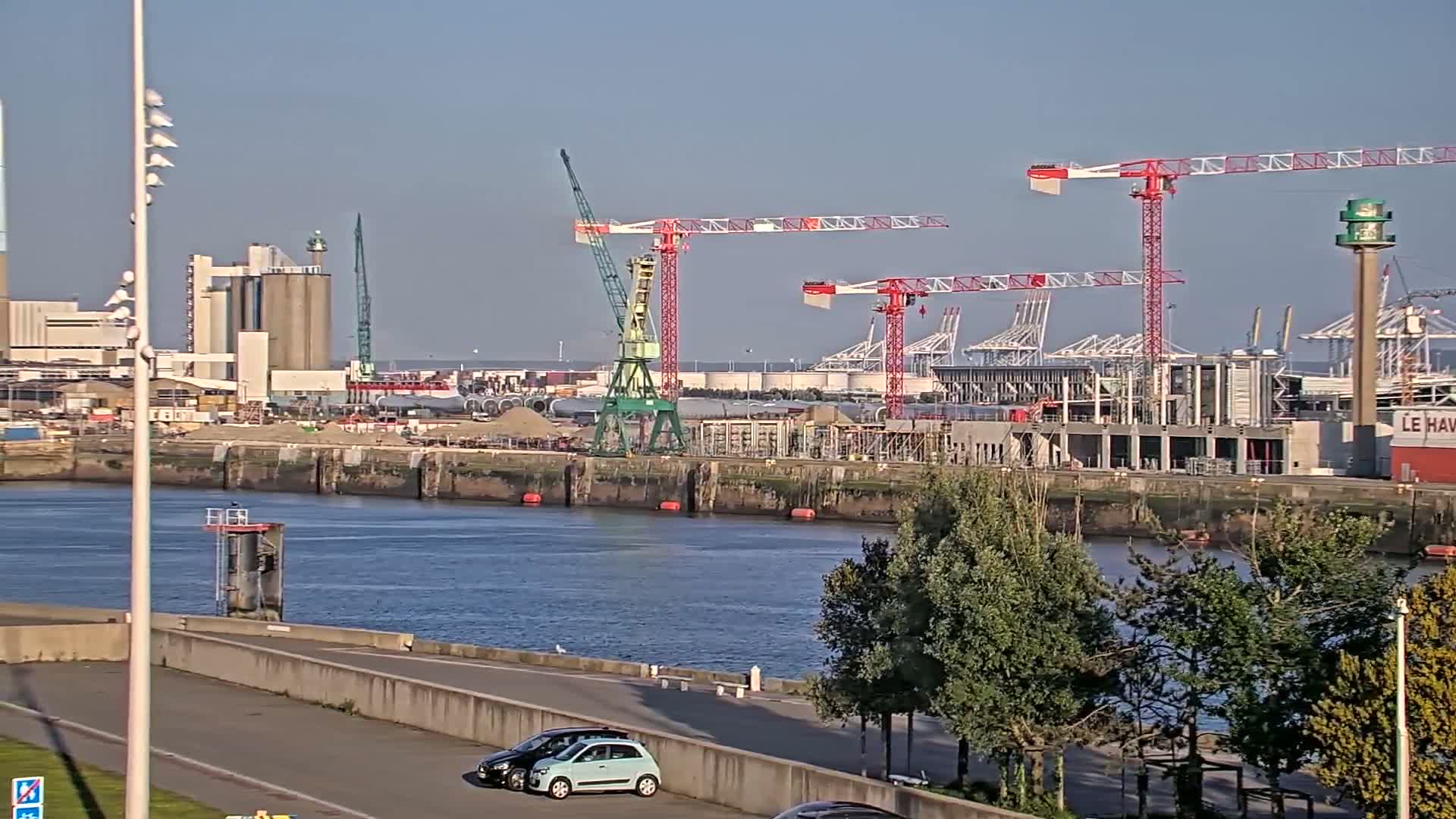 A waterfront view on a clear day shows a harbor with several cranes, industrial buildings, and construction activity, all next to a calm body of water with green trees along the shore.