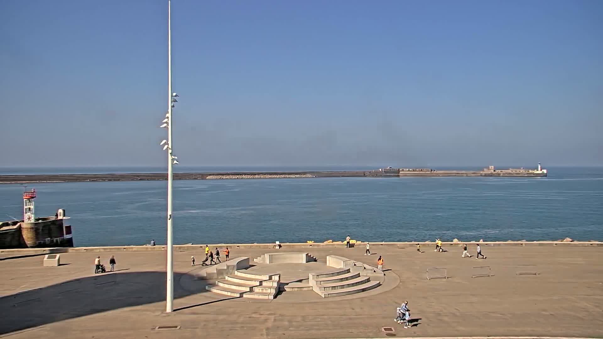 A paved waterfront area with a circular amphitheater-like structure, a tall flagpole, and several people, overlooks a calm ocean with breakwaters and a lighthouse in the distance under a clear blue sky.