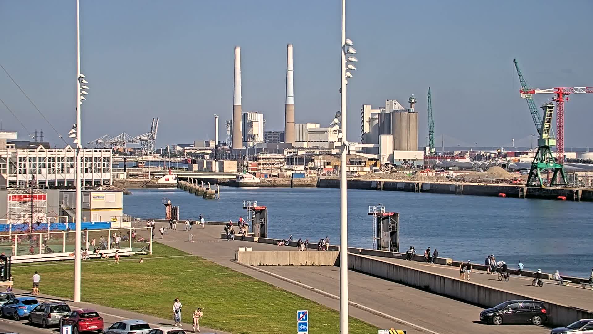 A sunny day at a harbor shows people walking and cycling along a paved waterfront path beside a grassy area, with an industrial complex including tall smokestacks visible across the water.