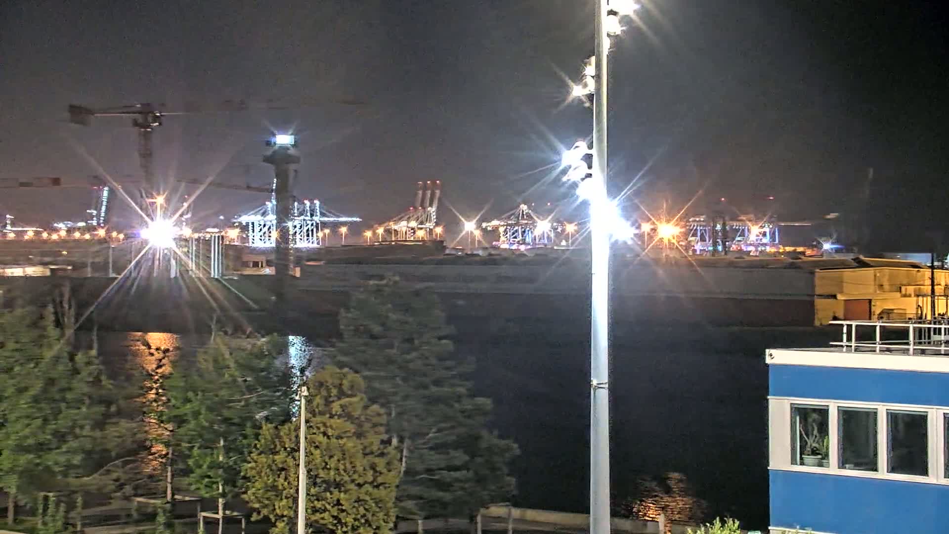 At night, a brightly lit industrial port area with cranes and ships is visible across a body of dark water from a vantage point with trees and a blue building in the foreground.