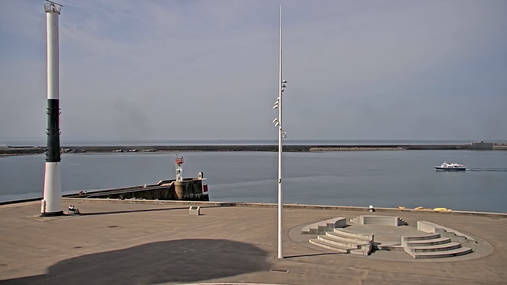 A harbor scene on a sunny day shows a tall white and black tower, a small lighthouse, an amphitheater-like structure, and a boat in the distance.