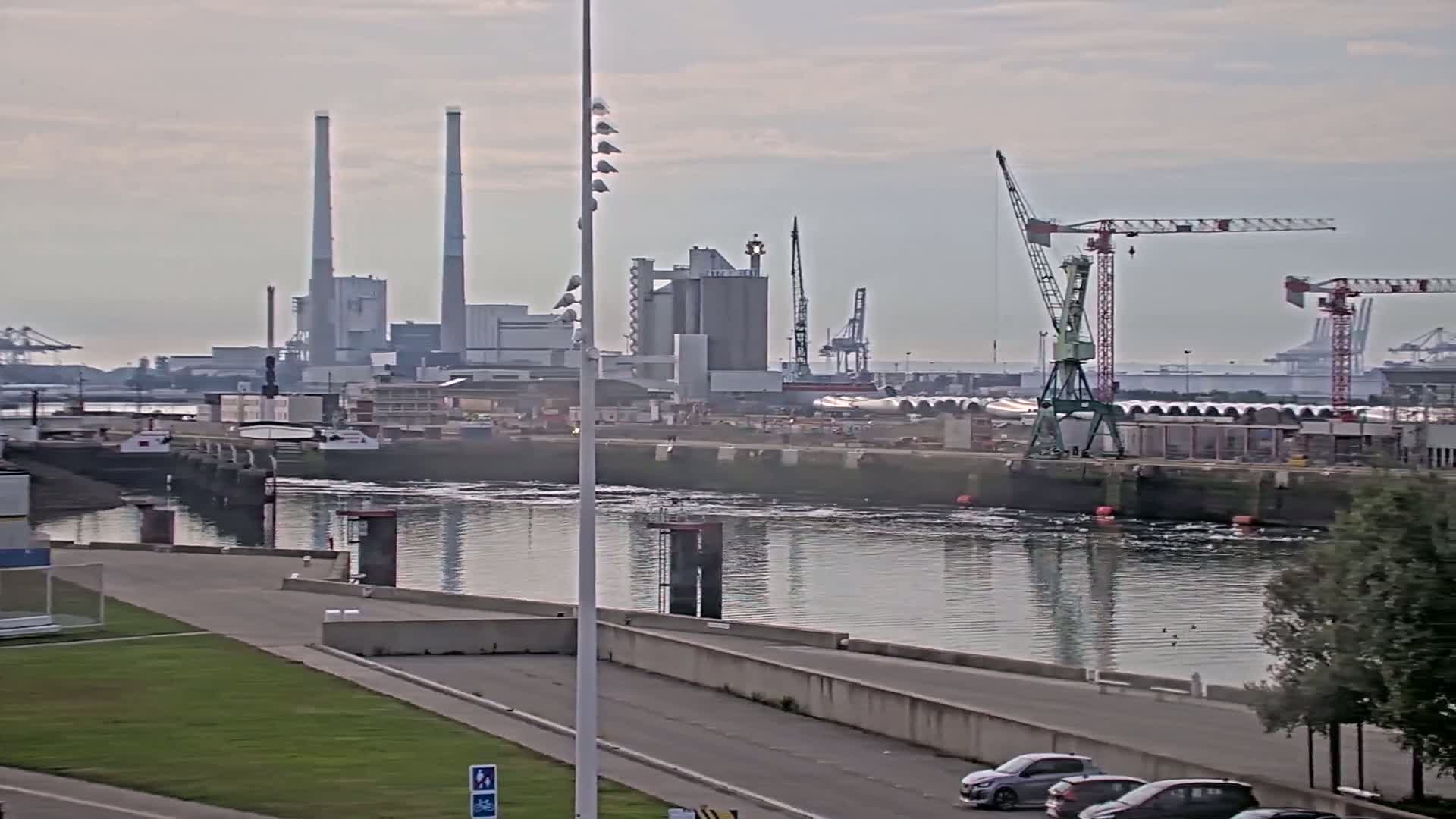 A calm body of water reflects an industrial waterfront scene under an overcast sky, featuring factories, cranes, and several parked cars along a paved road.