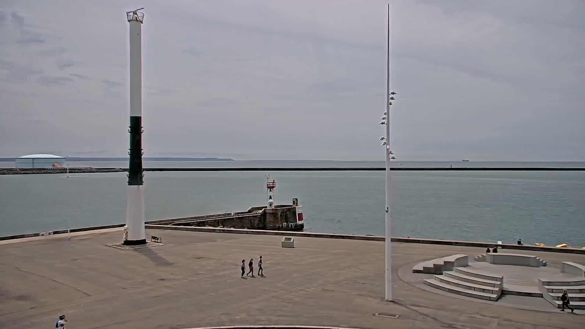 A mostly overcast day reveals a seaside paved area with two tall poles, a small stone amphitheater, and a few people walking near the calm water and breakwater.