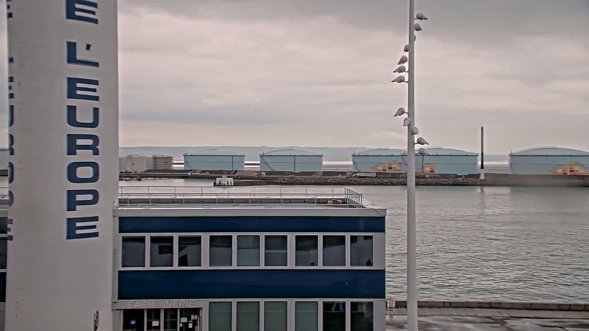 A blue building with a flat roof stands near a body of water under an overcast sky, with several large storage tanks visible in the distance.