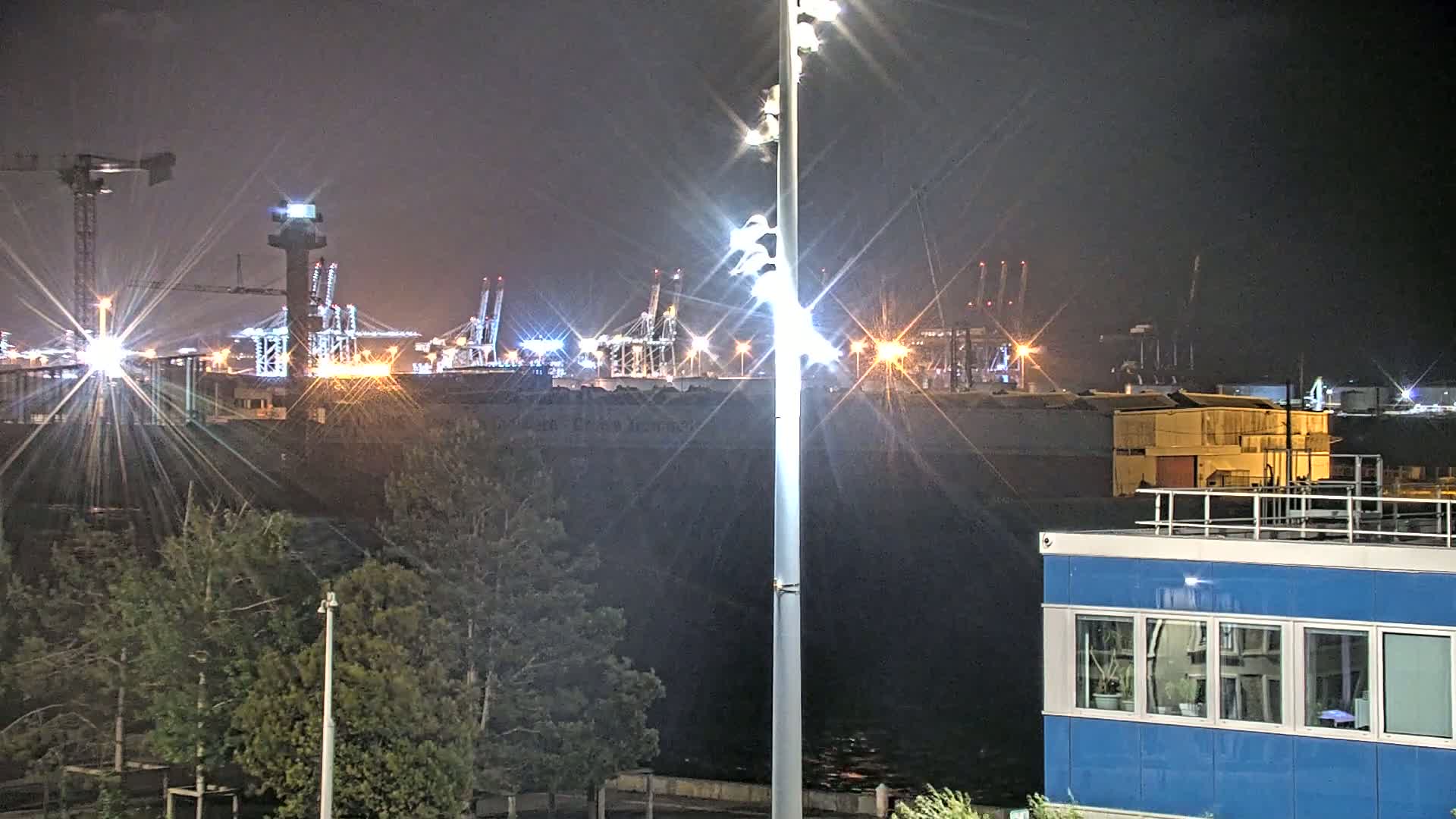 A brightly lit industrial port at night, viewed from across a body of water, with a tall light post in the foreground and trees in the mid-ground.