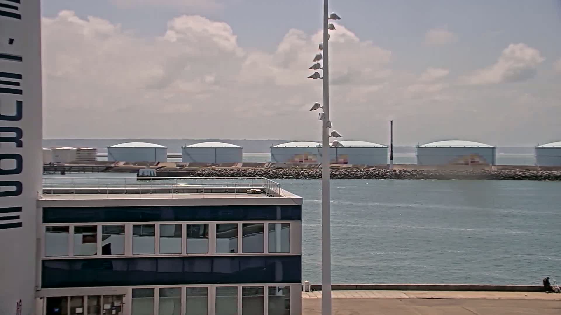 A partly cloudy day shows a body of water with several large, light-blue storage tanks across the water from a modern building with many windows.