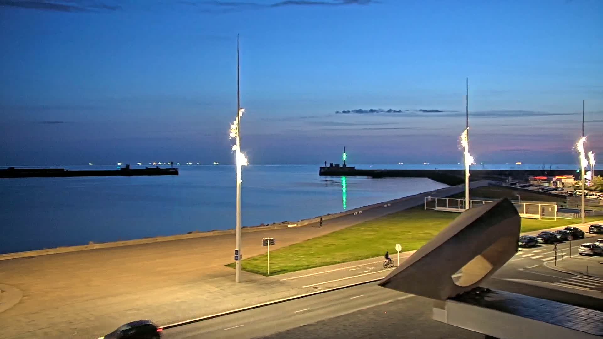 A twilight view of a calm harbor with lit pathways and a modern sculpture, showing several boats in the distance.