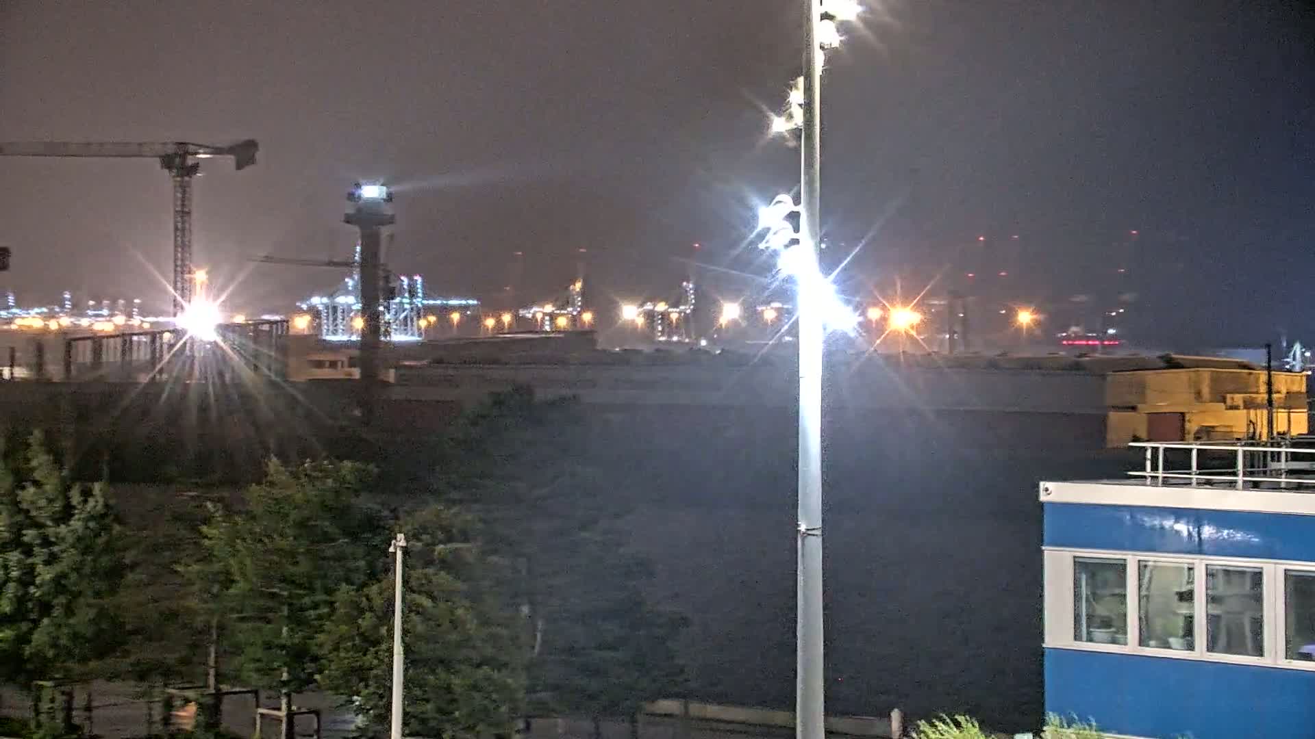 A nighttime view of an industrial waterfront, illuminated by bright lights, with rain and cranes visible in the distance.