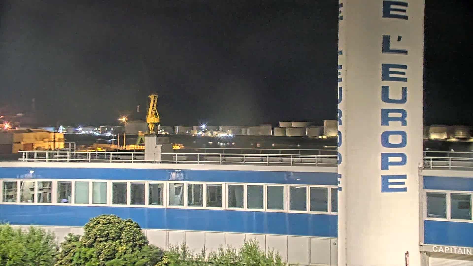 At night, a blue building with many windows is seen next to a tall column with "L'EUROPE" written on it, overlooking an industrial area with a crane and storage tanks.