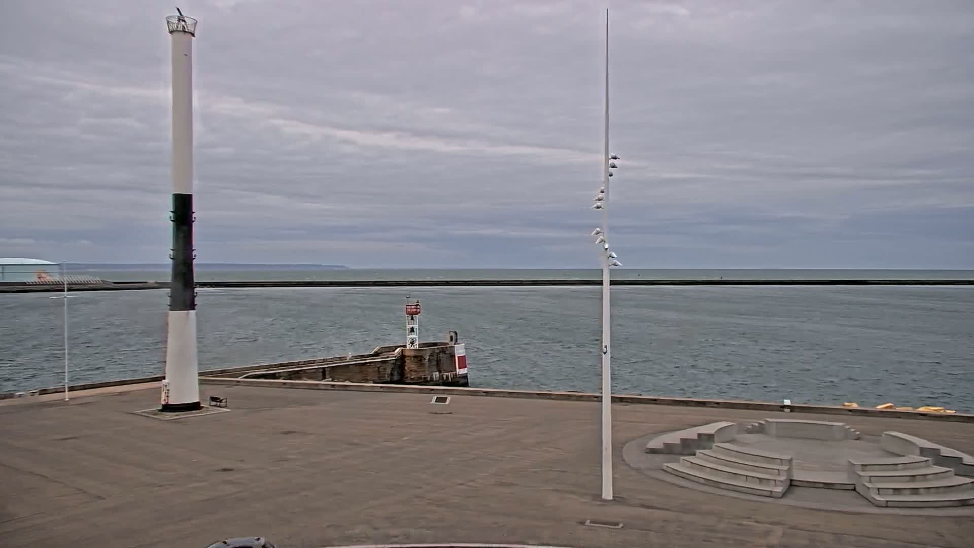 A harbor scene under a cloudy sky shows a tall, slender lighthouse, a smaller lighthouse on a breakwater, and an empty amphitheater-like structure in the foreground.