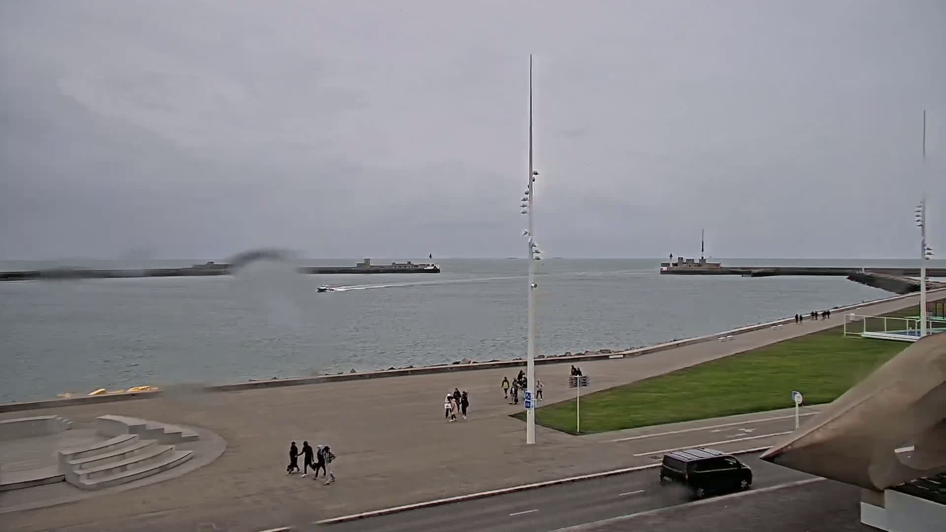 A calm, overcast day shows a harbor with a few people walking along a paved waterfront path, a small boat on the water, and a dark-colored vehicle driving on a nearby road.