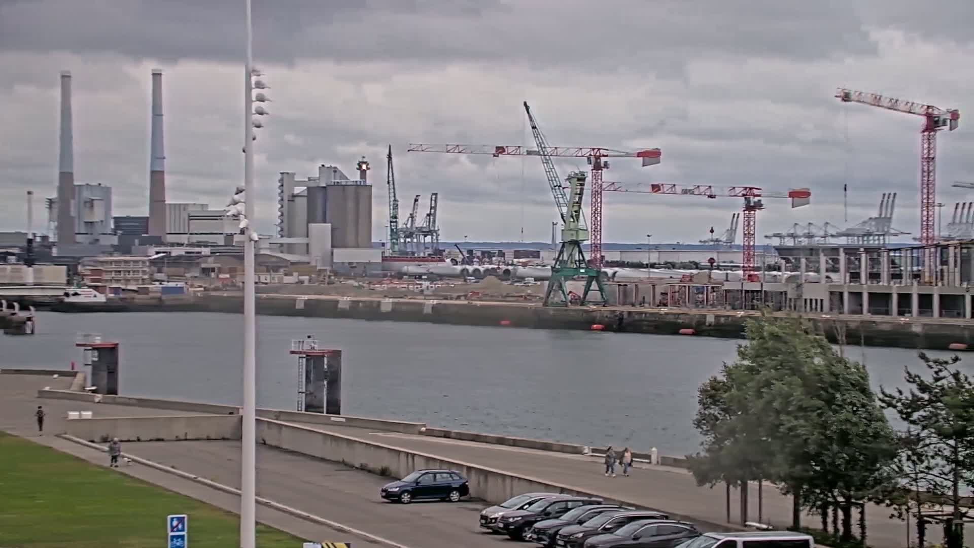 A waterfront industrial area with several cranes and factories under an overcast sky, showing a calm body of water and a paved walkway with a few people.