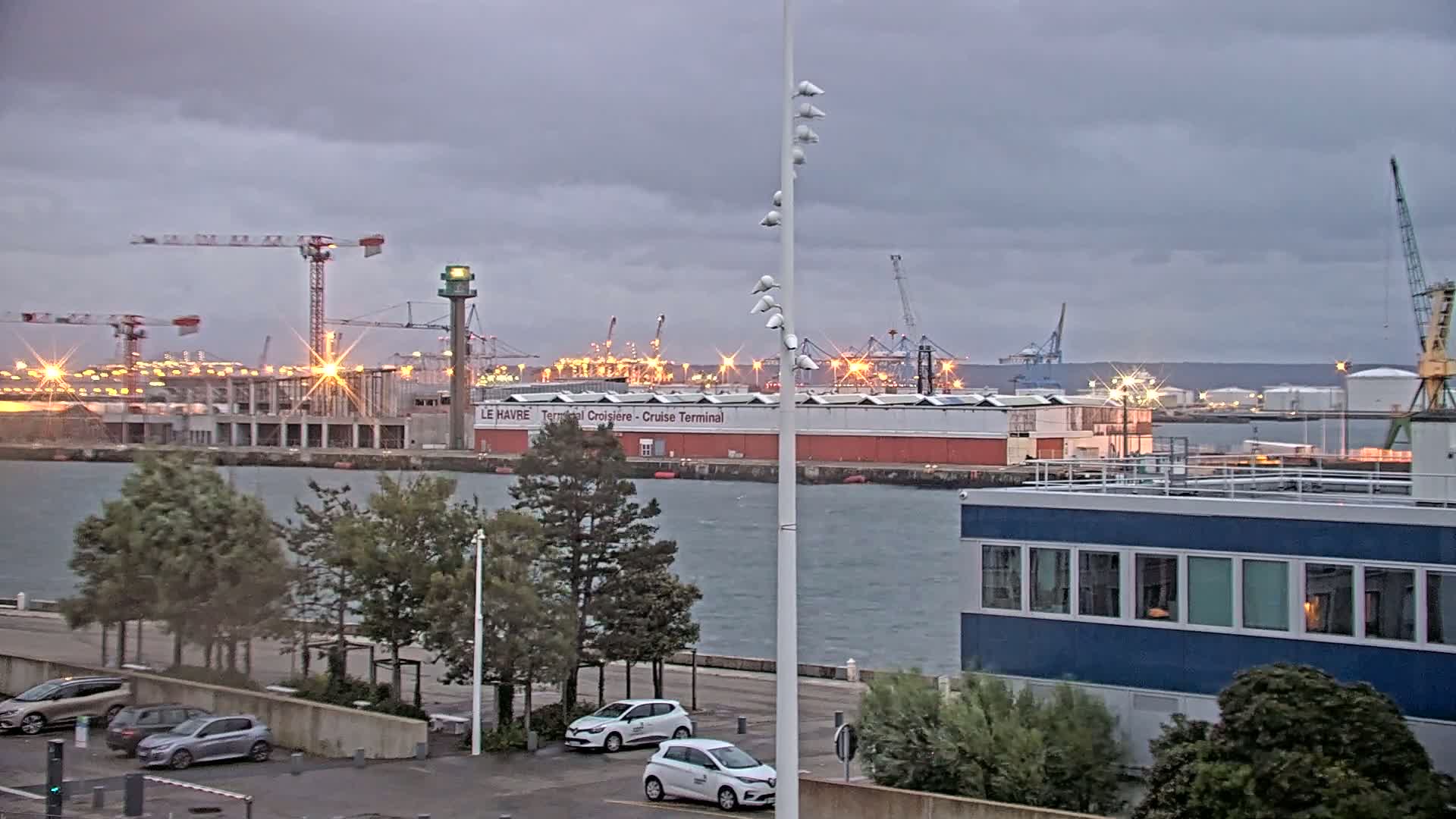 A harbor scene under an overcast sky shows a cruise terminal, construction cranes, several parked cars, and a blue building near the water.