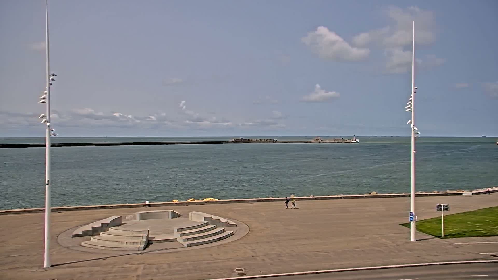 A mostly sunny day shows a paved waterfront area with a circular stone amphitheater and two tall light poles, overlooking calm ocean water and a distant breakwater.
