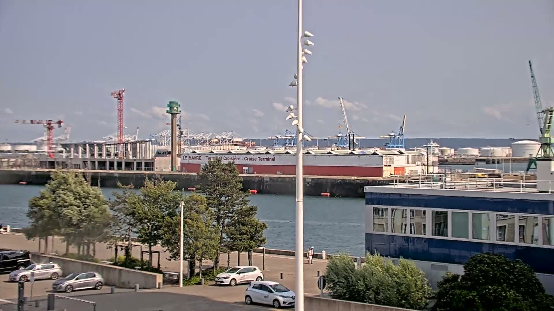 A harbor scene on a sunny day shows a large industrial area with cranes and buildings next to calm water, with several cars parked along a waterfront promenade.