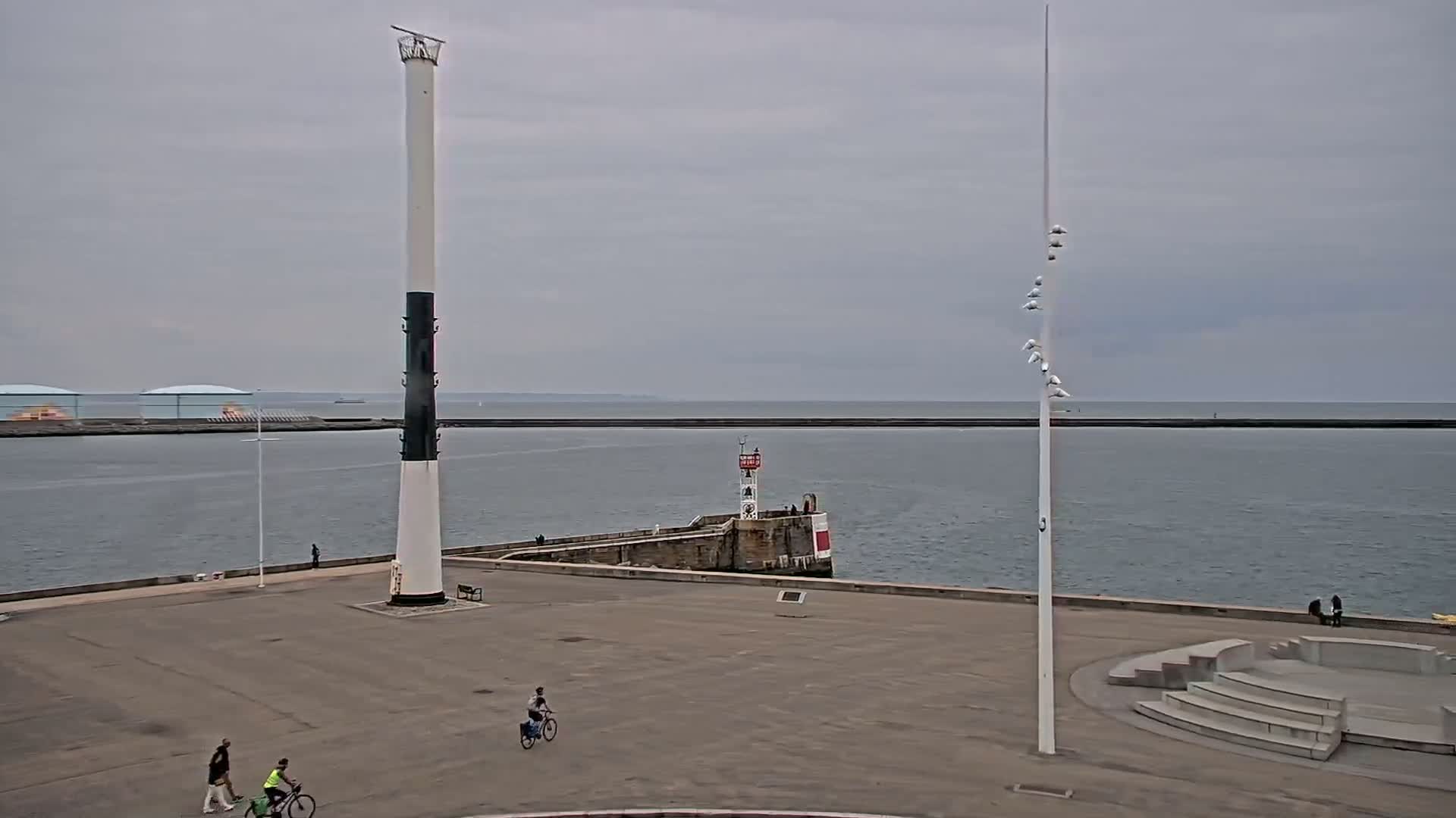 A mostly overcast day shows a harbor with a tall, black-and-white lighthouse, a smaller lighthouse on a breakwater, several people walking and bicycling on a paved area, and a seawall extending into a calm body of water.