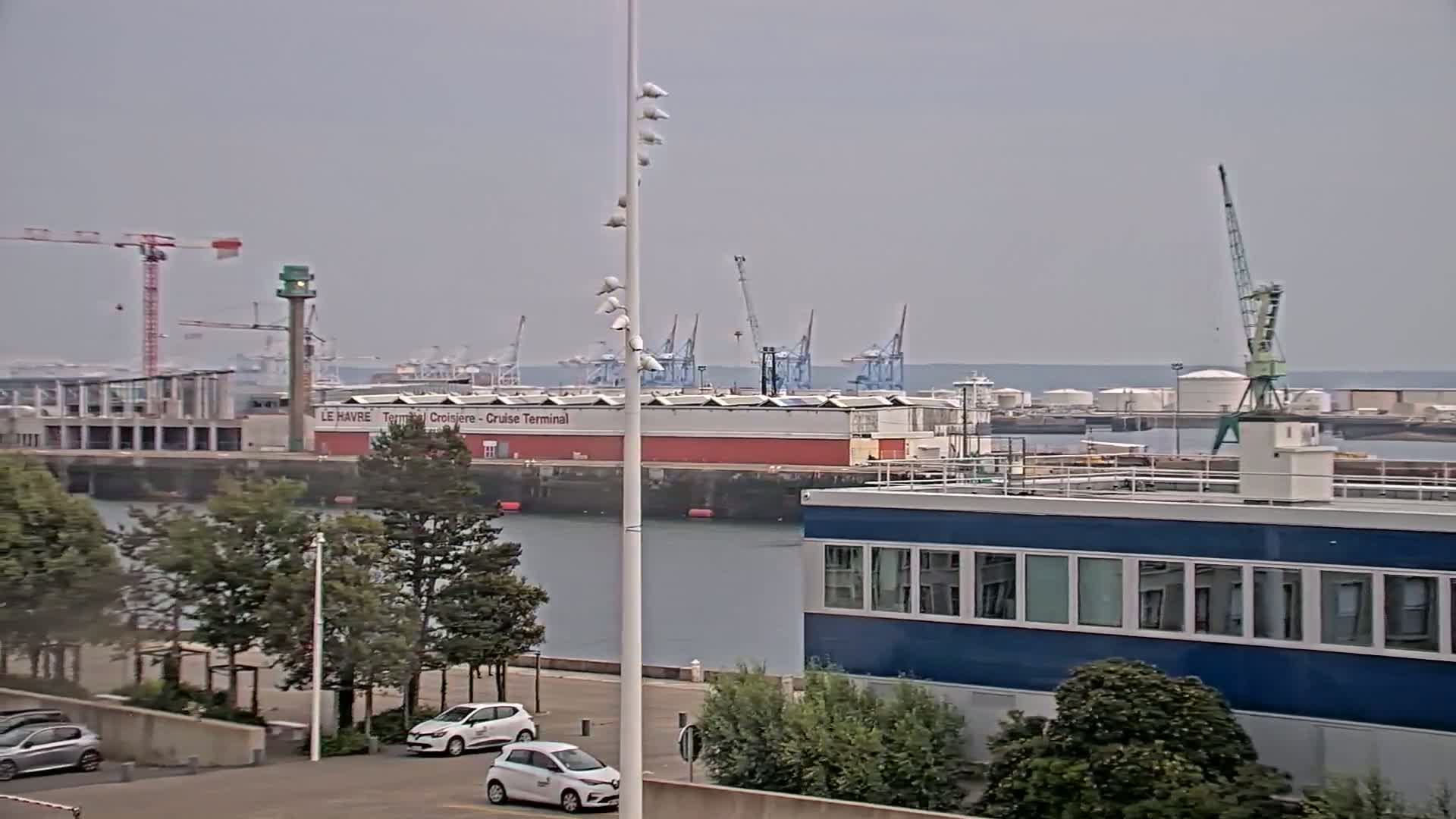 A harbor scene on an overcast day shows a cruise terminal, several cranes, and a blue building, with cars parked nearby and trees lining the waterfront.