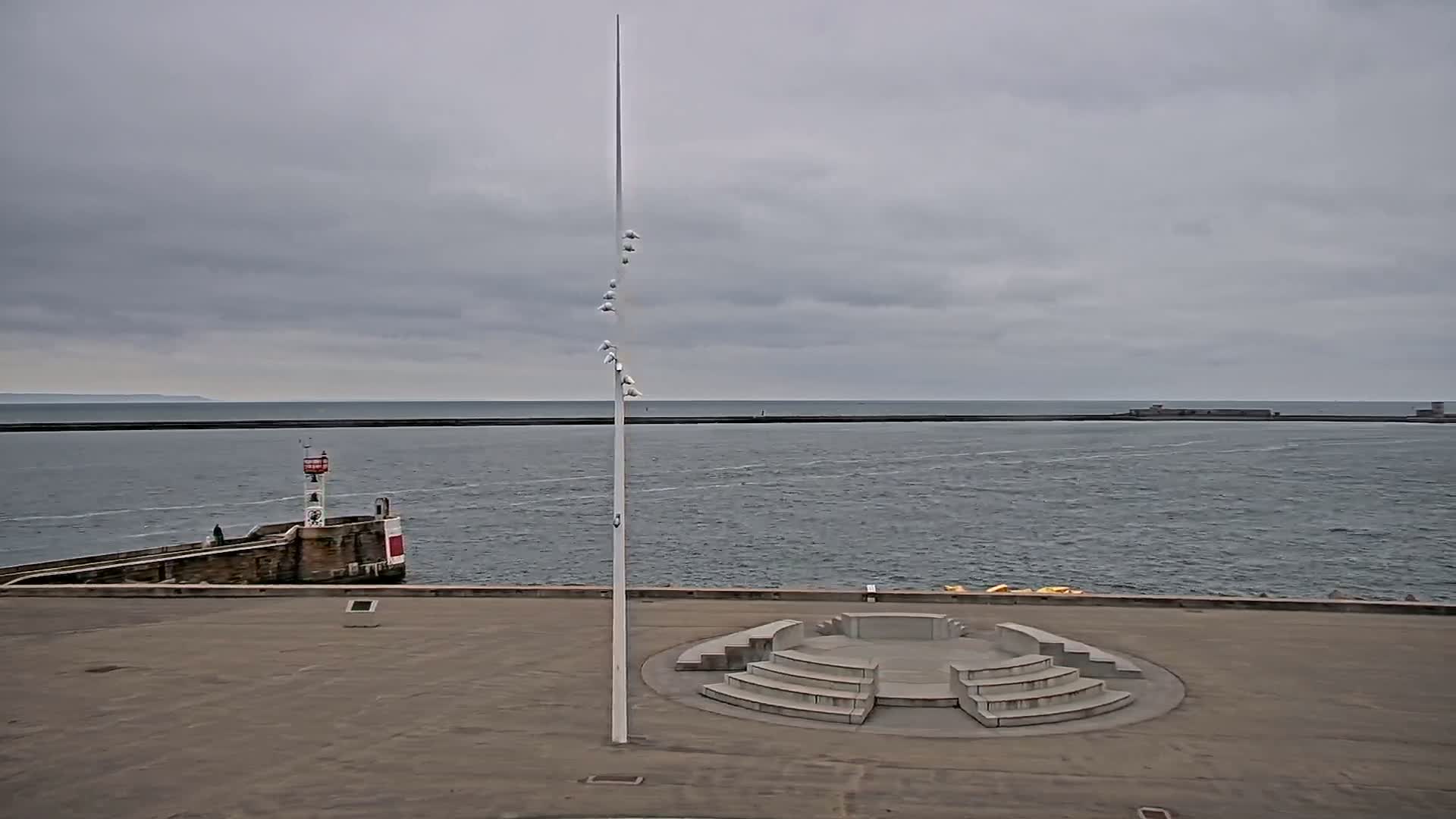 A concrete amphitheater-like structure sits on a paved area overlooking a calm sea under a cloudy sky, with a tall pole and a small lighthouse visible in the distance.