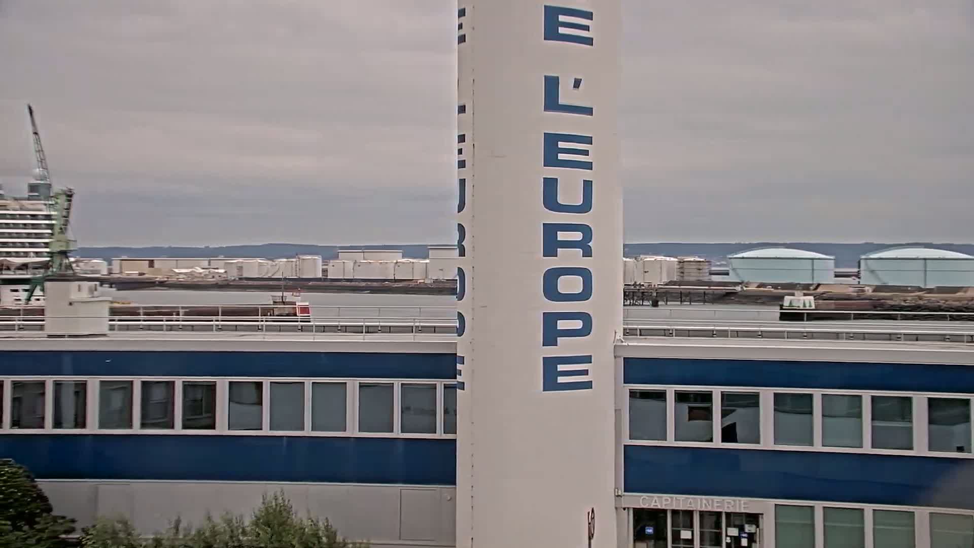 A tall white tower with "L'Europe" written on it stands in front of a blue building overlooking a body of water with industrial structures and storage tanks under an overcast sky.