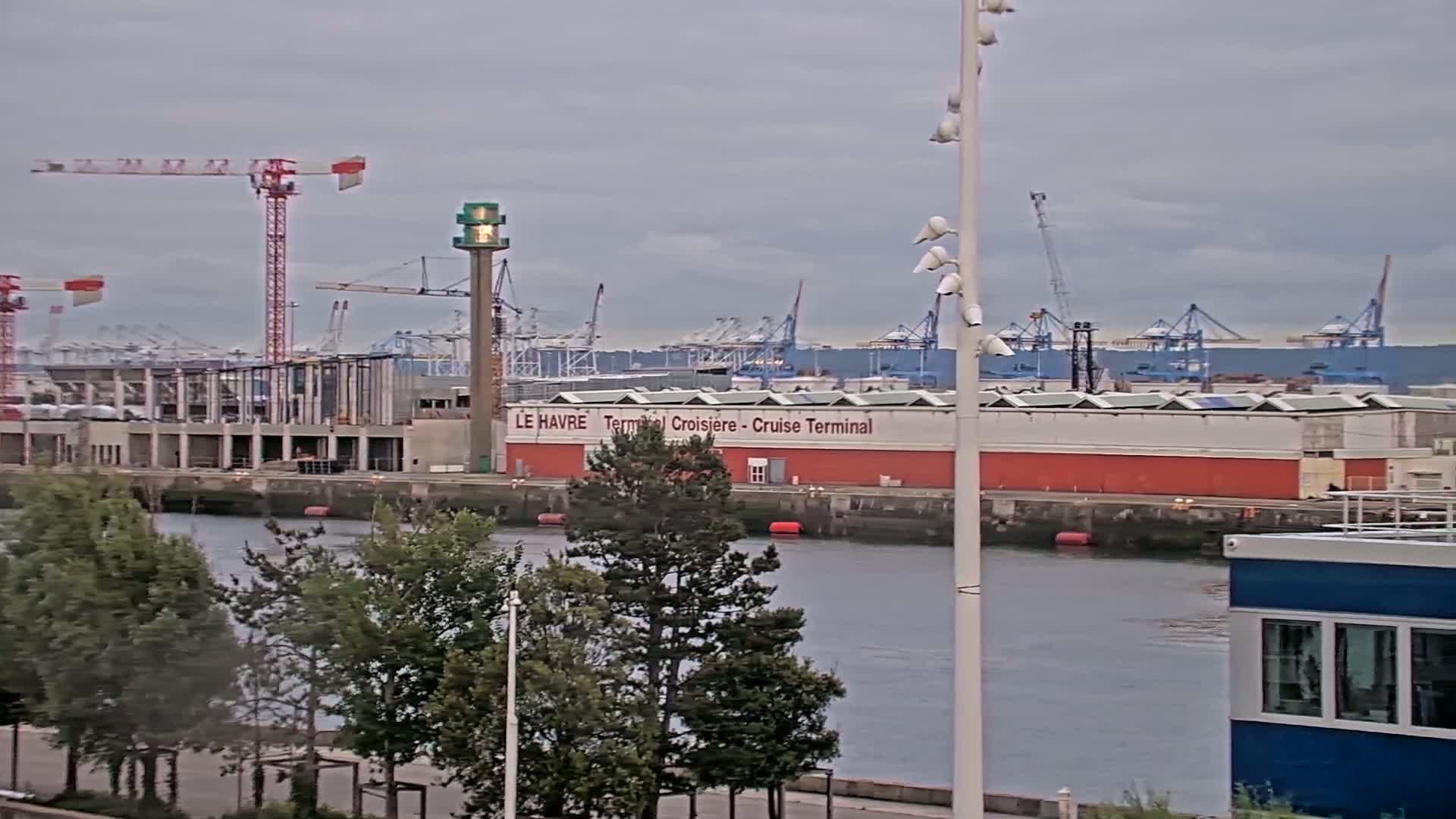 A harbor scene under overcast skies shows a cruise terminal building, construction cranes, and numerous cargo cranes in the background, with trees and a small blue building in the foreground.