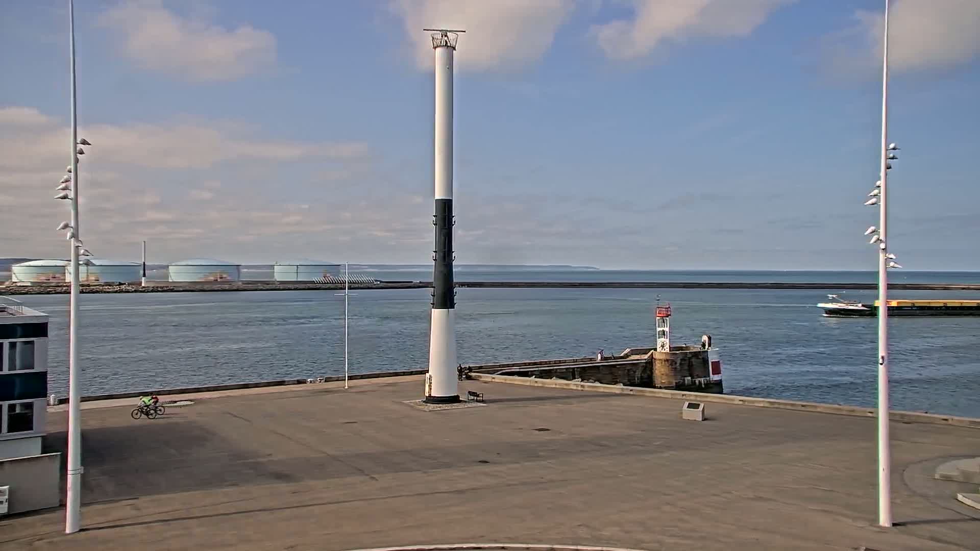 A tall, black-and-white lighthouse stands in a harbor on a partly cloudy day, with a cyclist on a paved area and a barge in the distance.