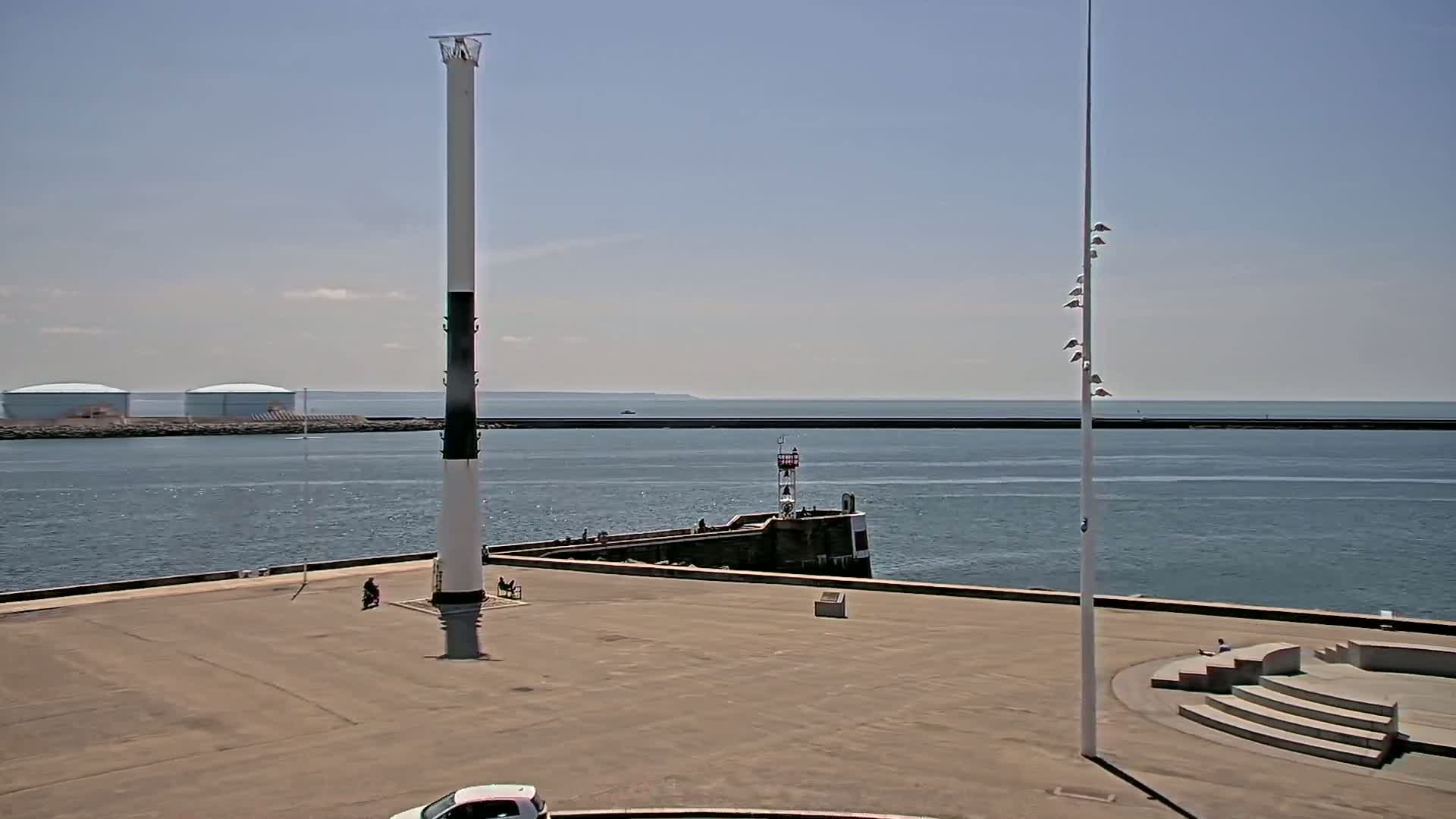 A mostly sunny day at a harbor shows a tall, black and white striped pole, a small lighthouse, and a wide expanse of calm water with a distant breakwater and industrial storage tanks.