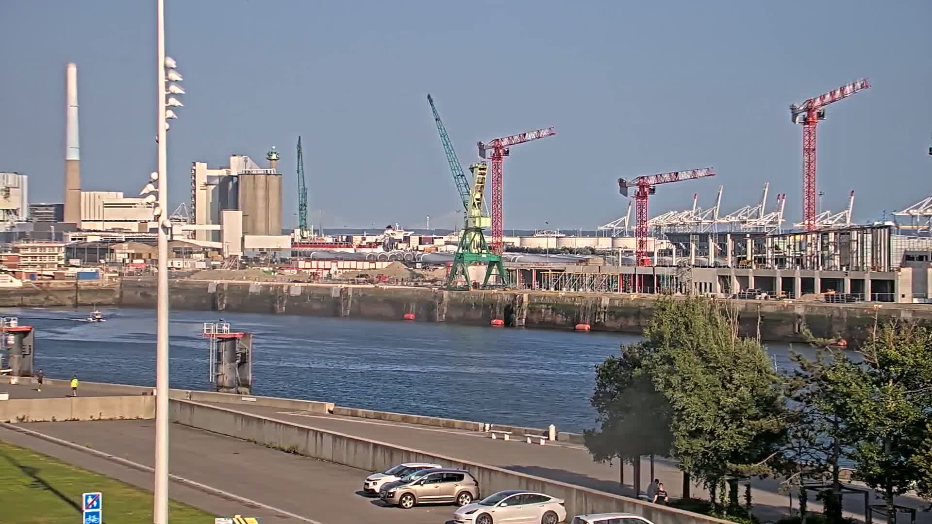 A sunny day at a harbor shows several cranes and industrial buildings under construction across a body of water, with a few cars parked along a paved waterfront path.