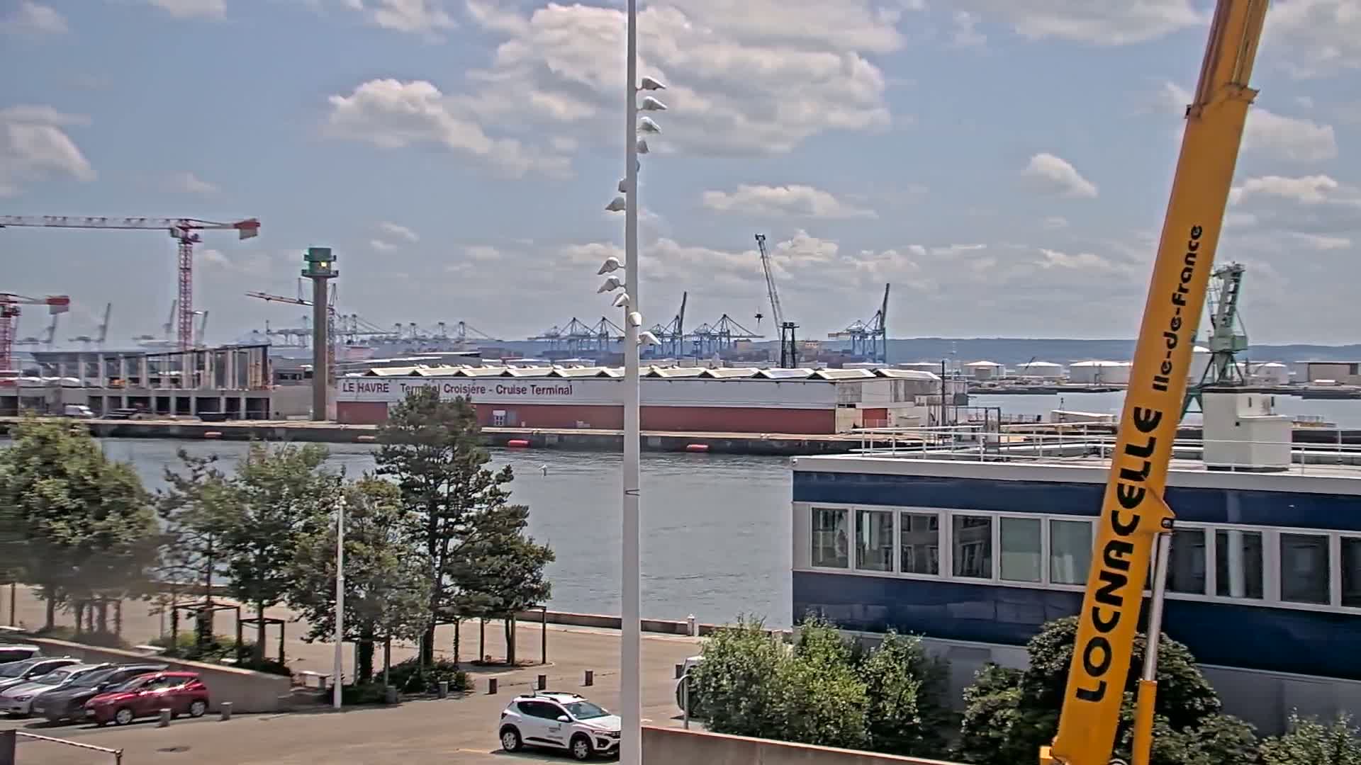 A partly cloudy day at a harbor shows a cruise terminal, several cranes, and docked ships; a large yellow crane is in the foreground.