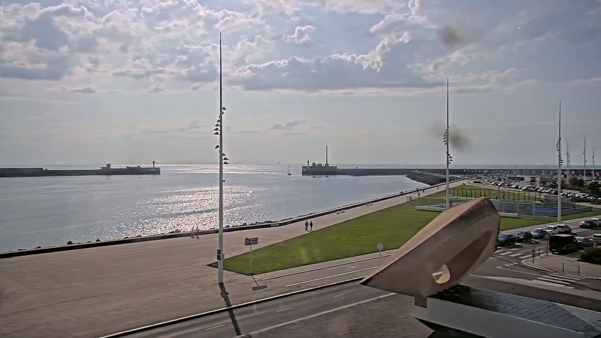 A partly sunny day reveals a harbor with calm water, a paved walkway, grassy areas, and parked cars, all situated near a large, abstract sculpture.