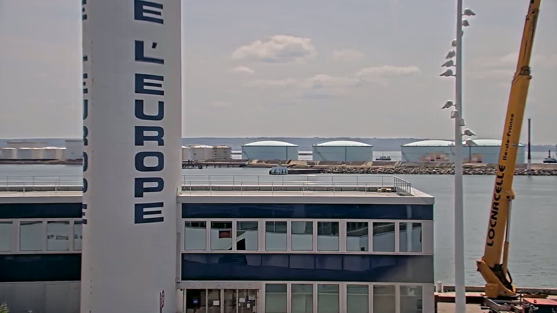 A tall white column with "L'EUROPE" written on it stands next to a building with many windows overlooking a body of water where several large storage tanks are visible in the distance under a partly cloudy sky.