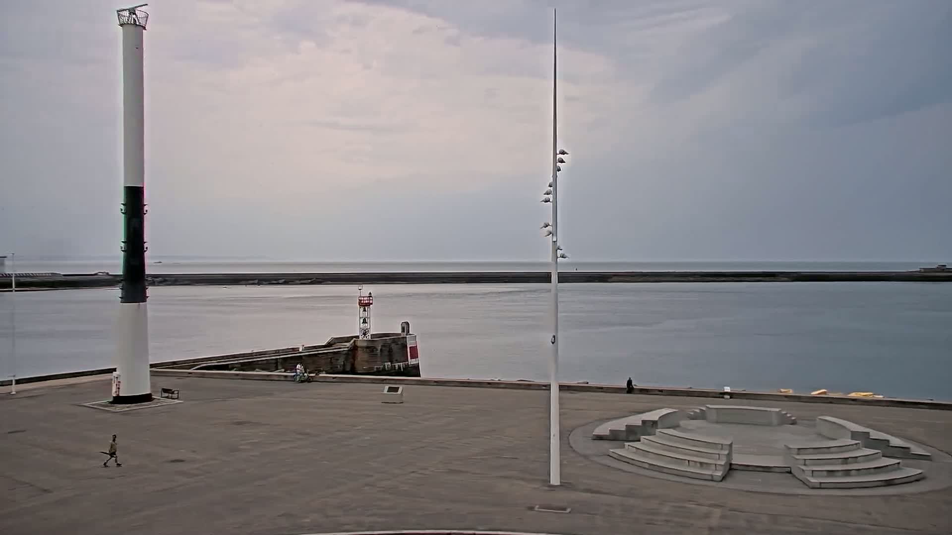 A harbor scene under a cloudy sky features a tall, slender lighthouse, a smaller lighthouse on a breakwater, and an amphitheater-like structure in a paved area overlooking calm water.