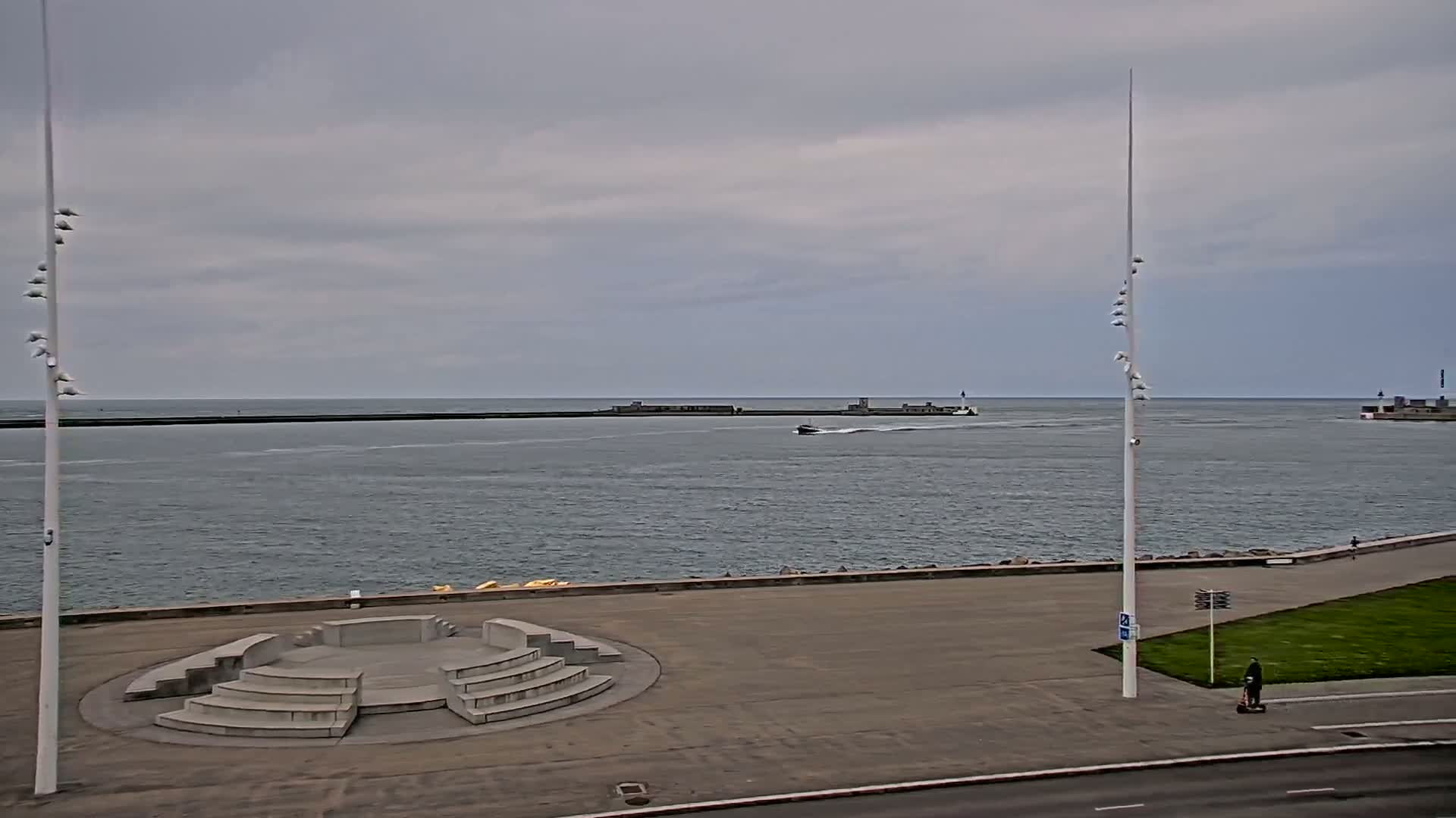 A mostly overcast day shows a harbor with a small boat and breakwaters, viewed from a paved waterfront area with a concrete amphitheater-like structure.