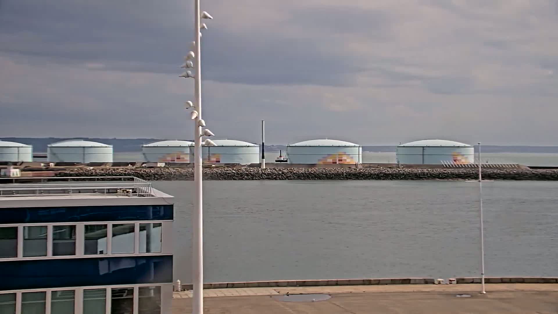 Several large, light-blue storage tanks are situated across a body of water from a modern building under a cloudy sky.