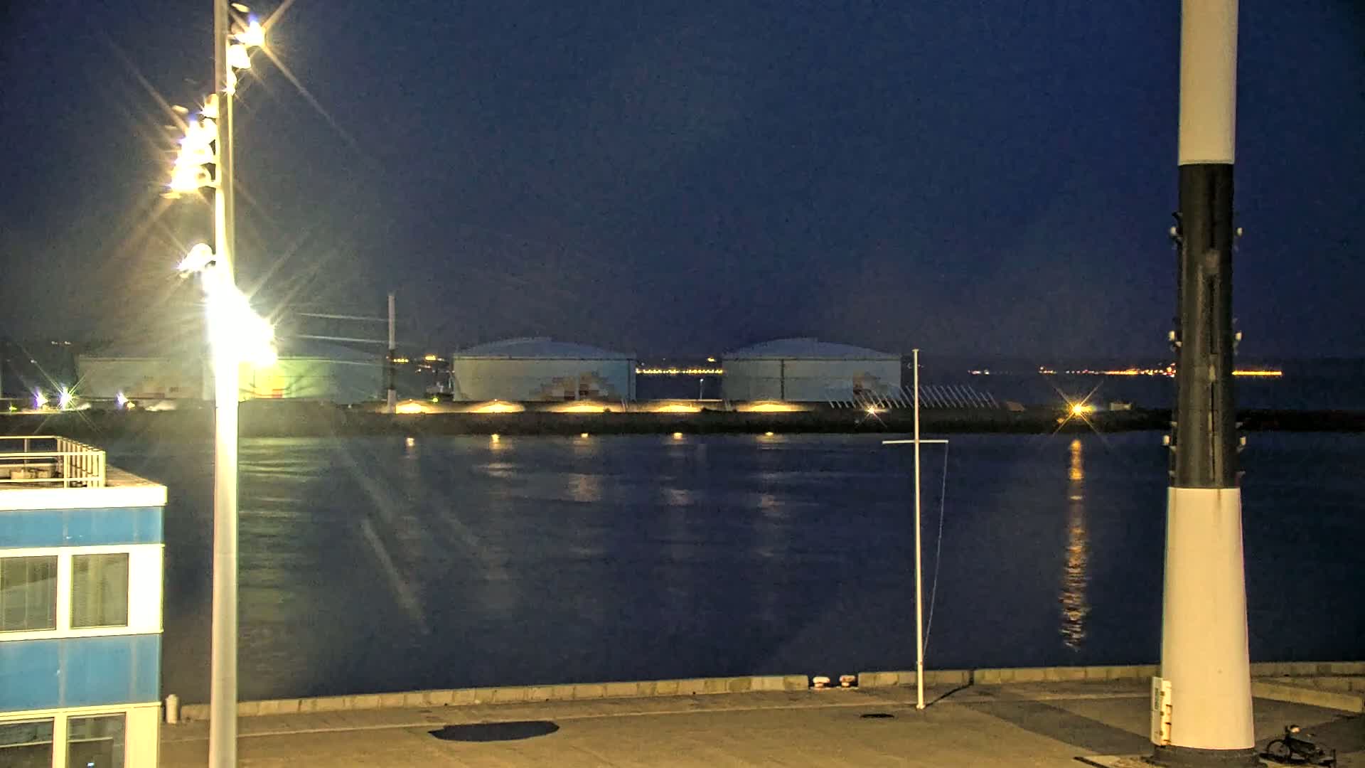 Several large, light-blue storage tanks are situated across a body of water from a modern building under a cloudy sky.
