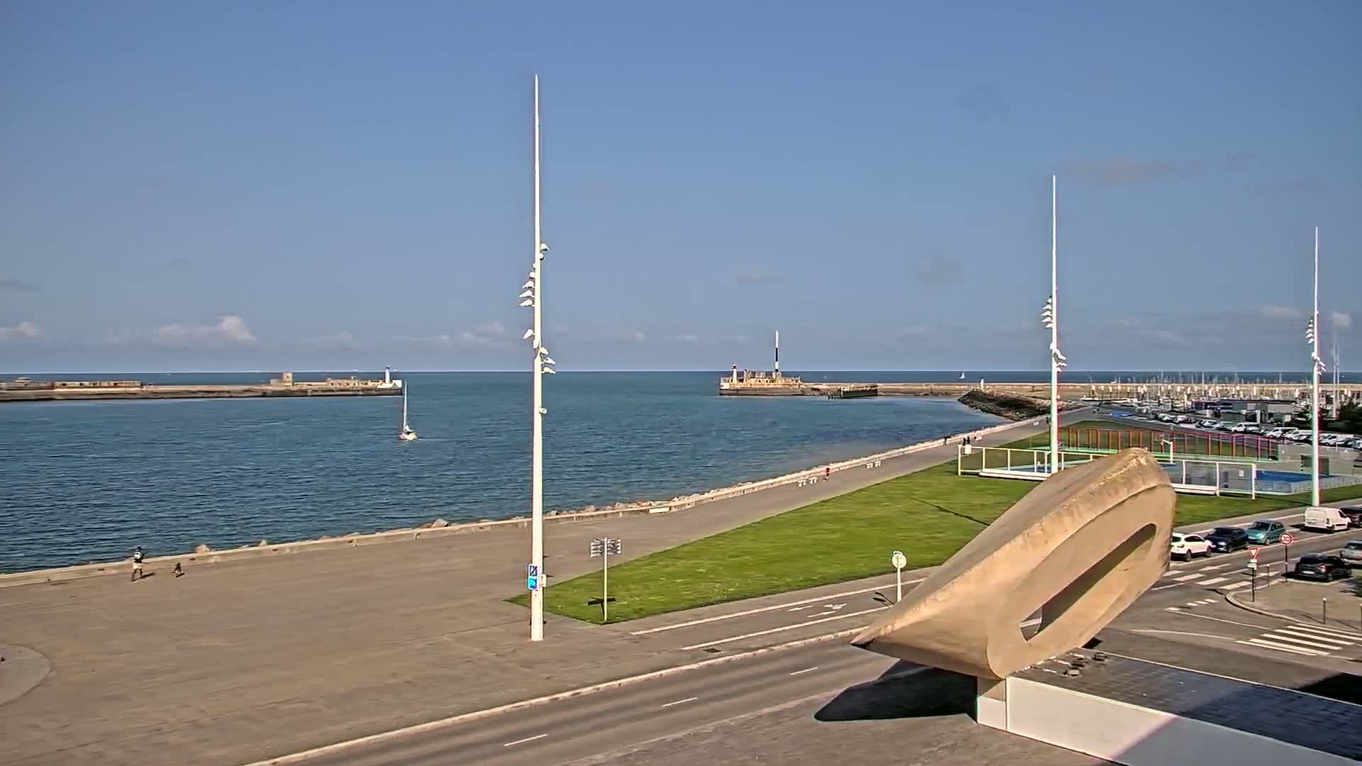 A harbor with calm water under a mostly clear blue sky, featuring a paved walkway, grassy areas, and a modern sculpture, with cars parked nearby.