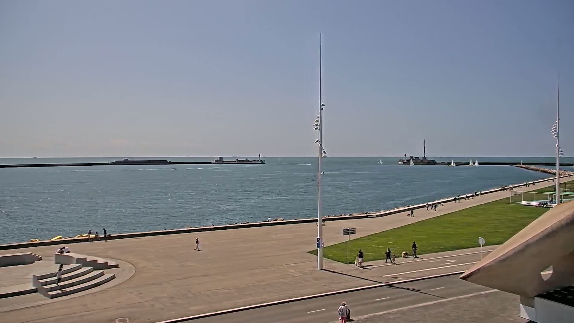 A harbor with a paved walkway and grassy areas, a few people strolling along the path, boats on the water under a clear sky.