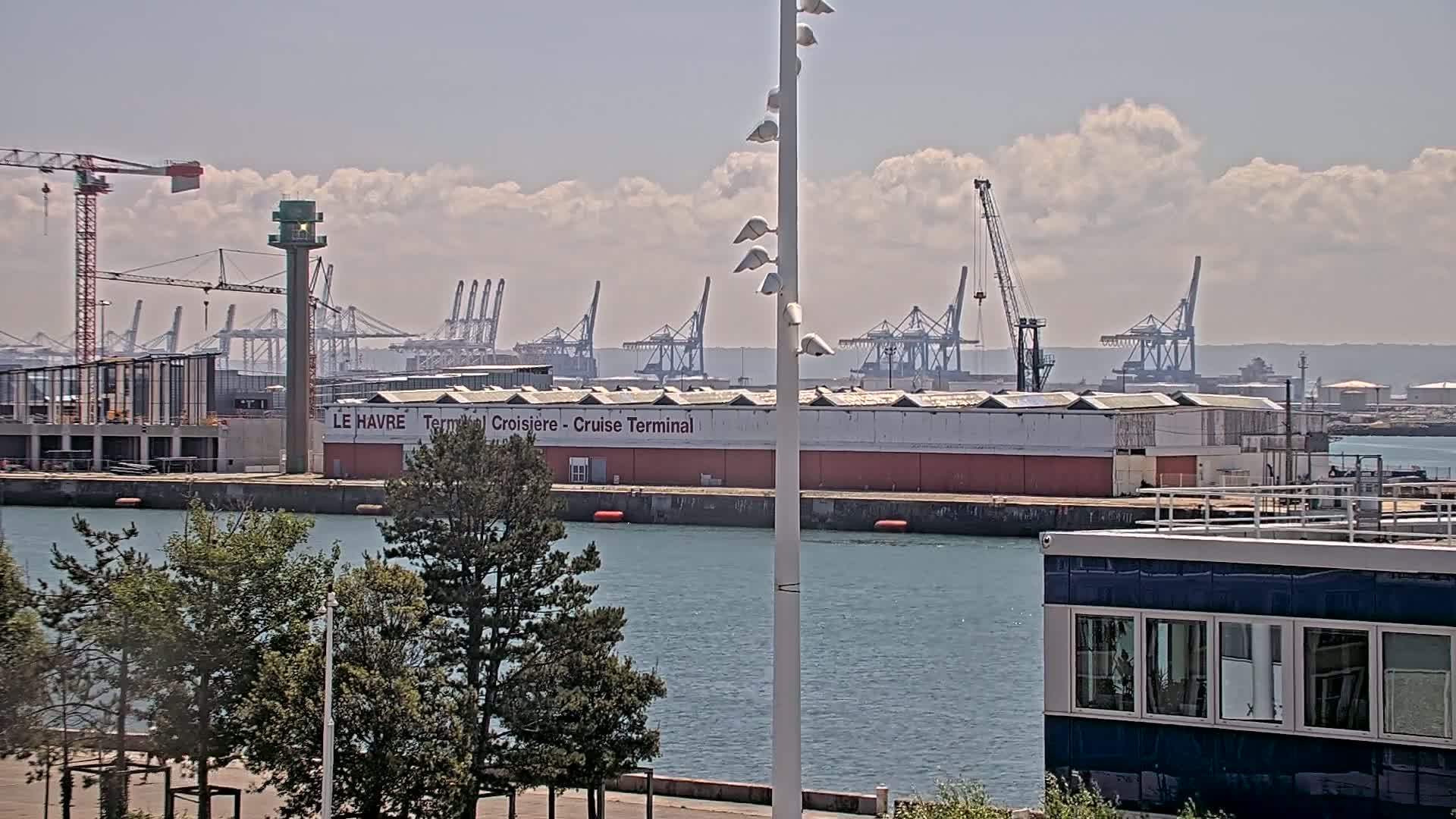 A harbor scene on a partly cloudy day shows a cruise terminal, numerous cranes, and other industrial buildings along a calm body of water, with green trees in the foreground.