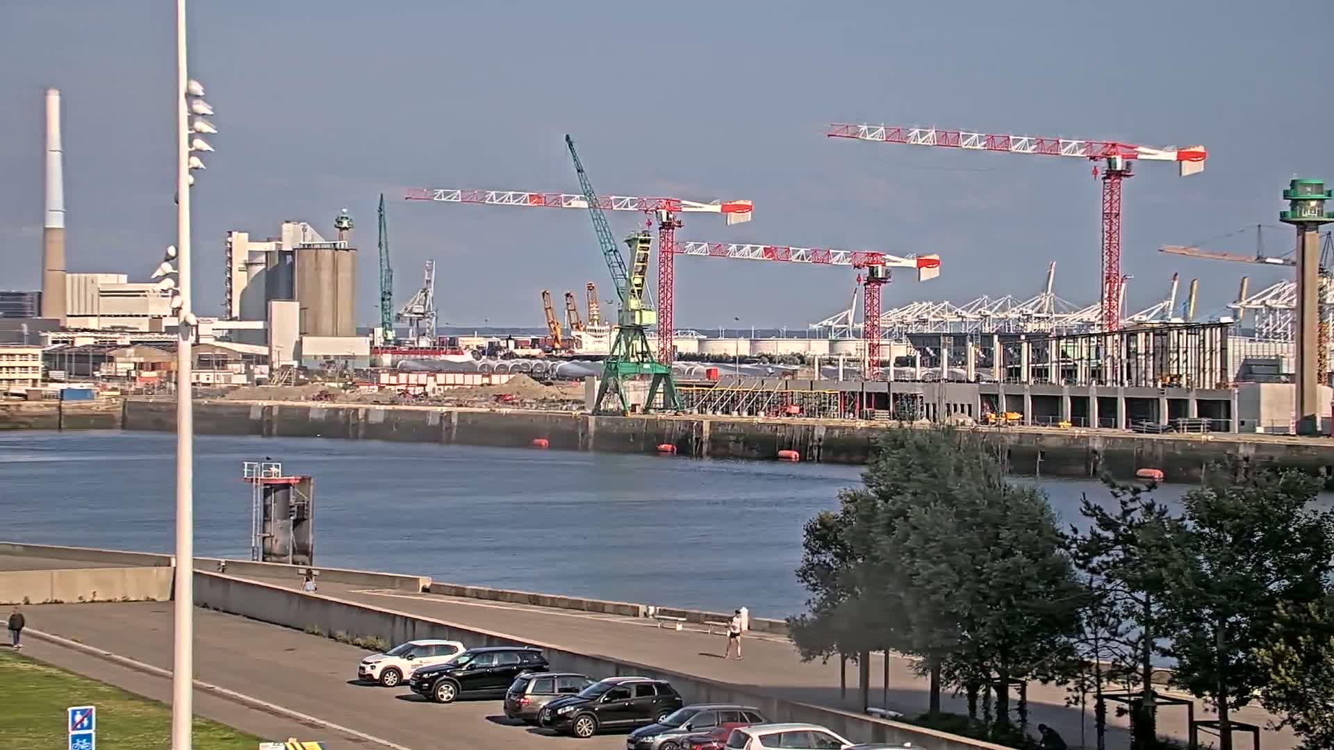 A waterfront construction site with several cranes is visible across a calm body of water on a sunny day.